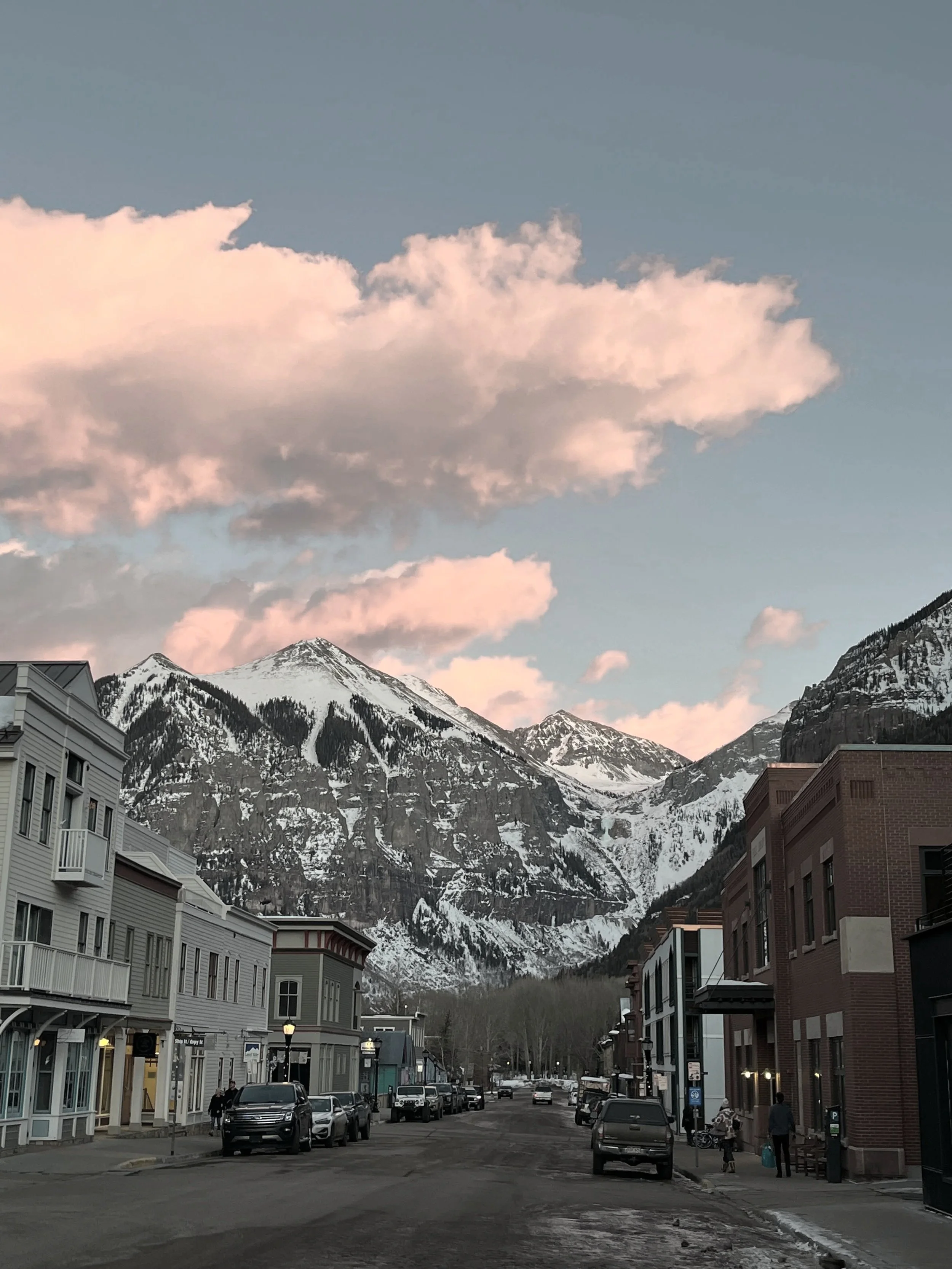 Street scene in a mountain town with snow-capped peaks in the background and pink clouds in the sky at sunset