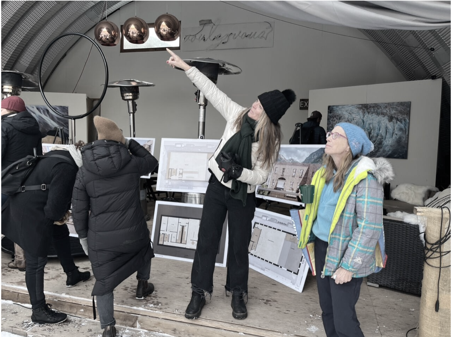 Group of people at a model display in a studio with architecture floor plans and landscape images, one woman pointing upward, dressed warmly with a hat and scarf, others examining plans.