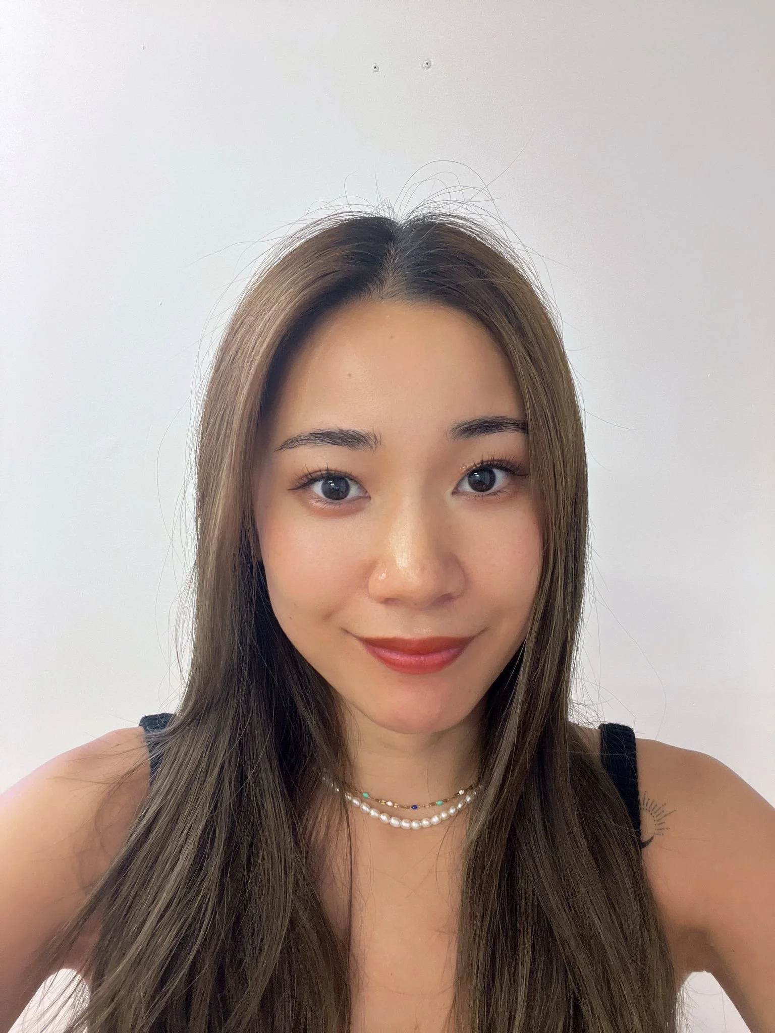 Young woman with long hair smiling, wearing a black top and necklaces, against a plain background.