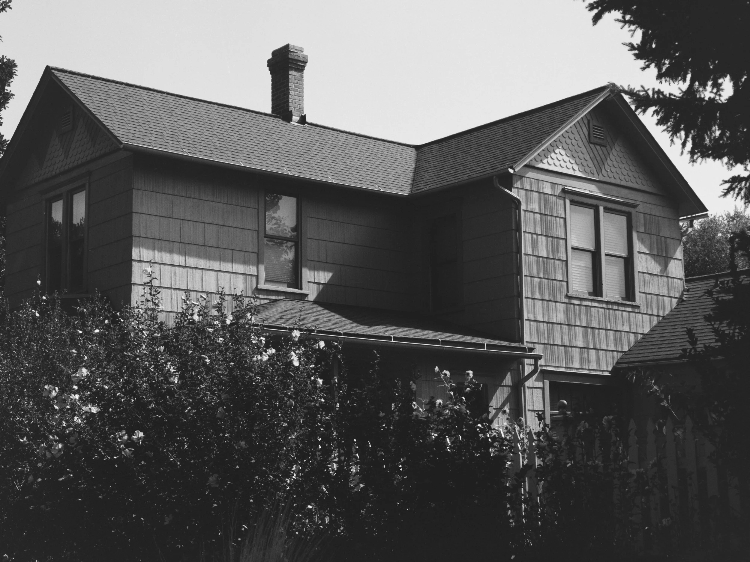 A two-story house with a chimney, windows, and a sloped roof, surrounded by trees and bushes, in black and white.