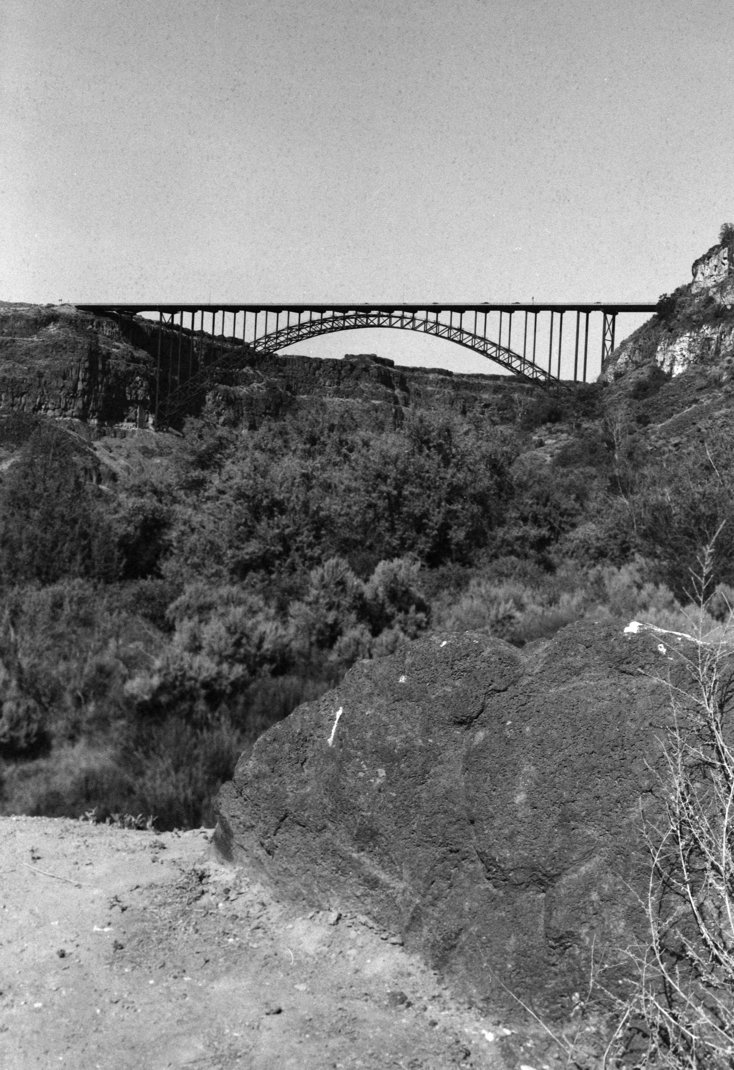 A black and white photo of a large arch bridge spanning across a deep canyon with rocky cliffs and dense vegetation below.