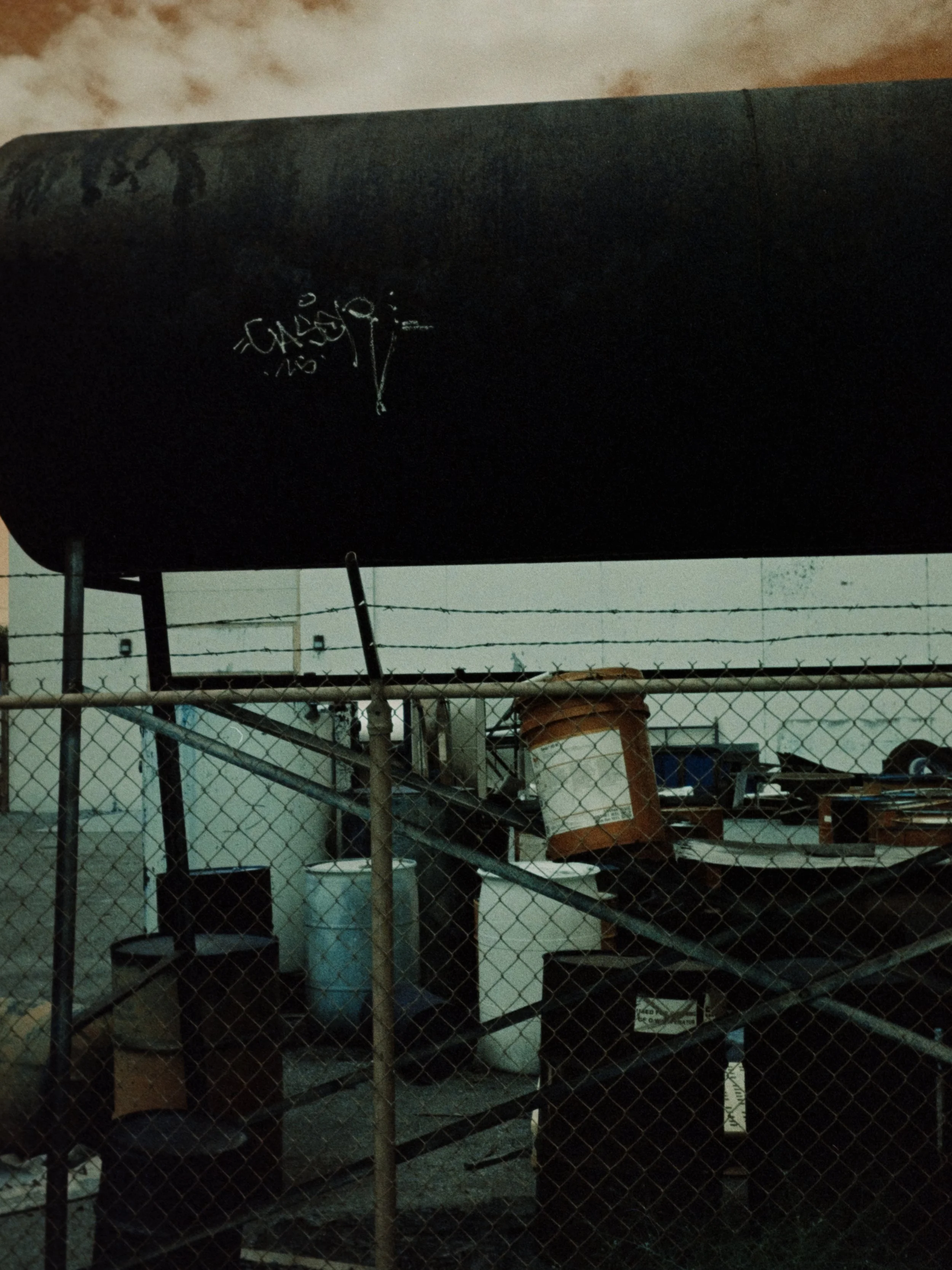 Storage yard with barrels and boxes behind a chain-link fence, above the yard is a large black fuel tank with graffiti.