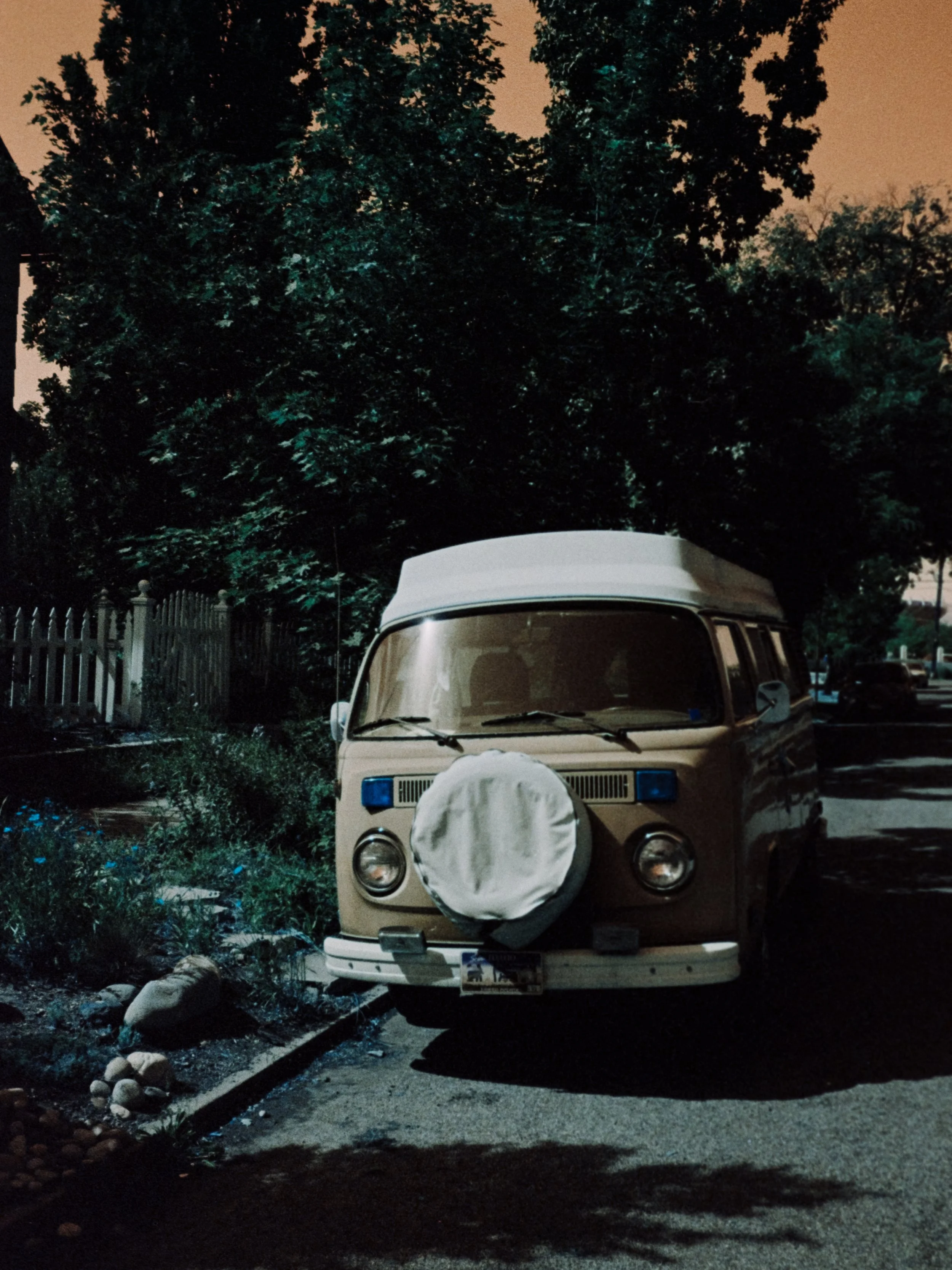 A vintage beige Volkswagen camper van parked on a street at night, with a large leafy tree behind it and a wooden fence to the left.