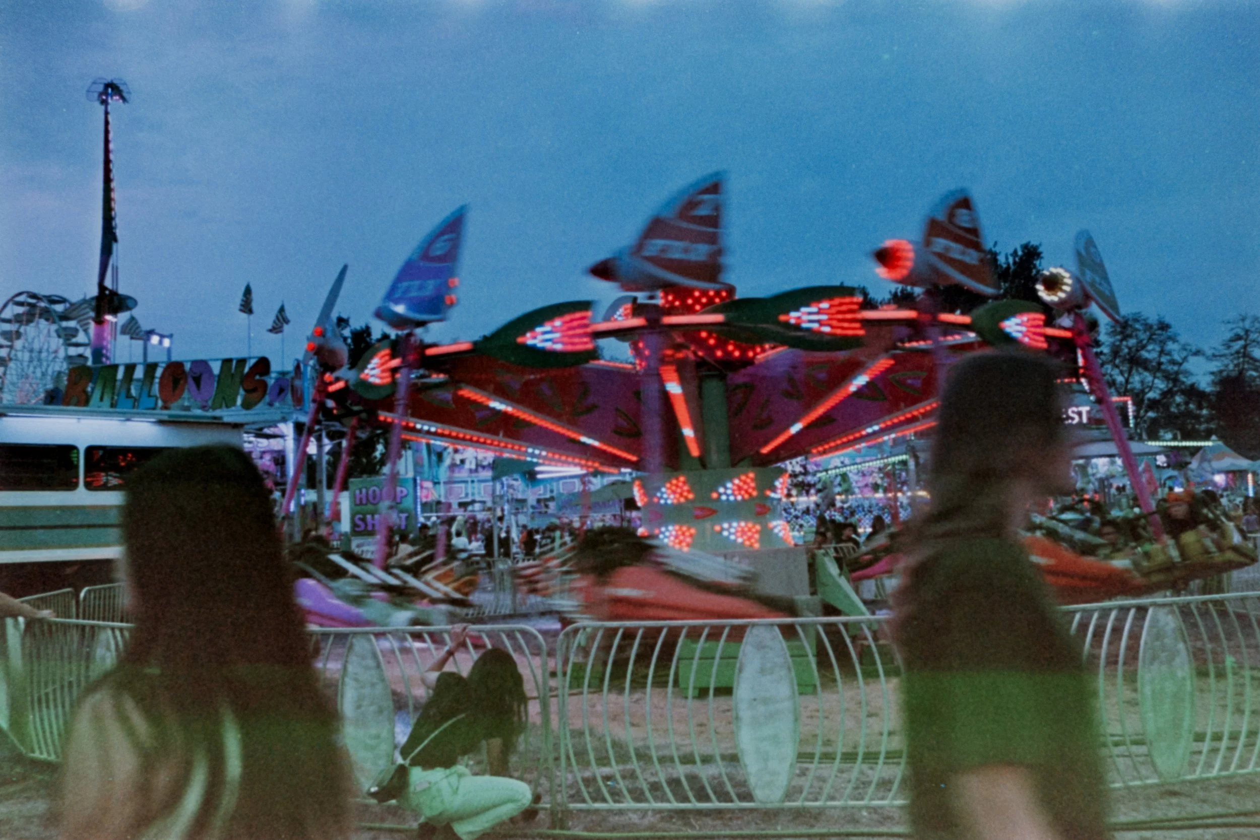 An amusement park at dusk featuring a brightly lit spinning ride with children on it, and a few people walking past in the foreground.