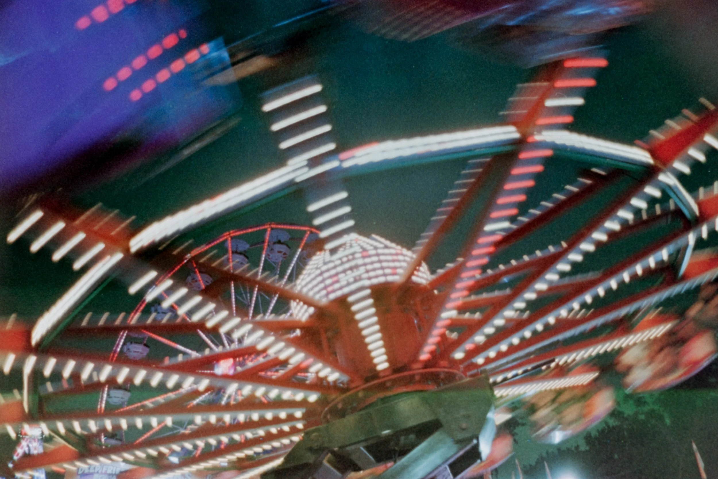 Long exposure photograph of a carnival ride with bright lights and spinning motion, showing a Ferris wheel in the background at night.