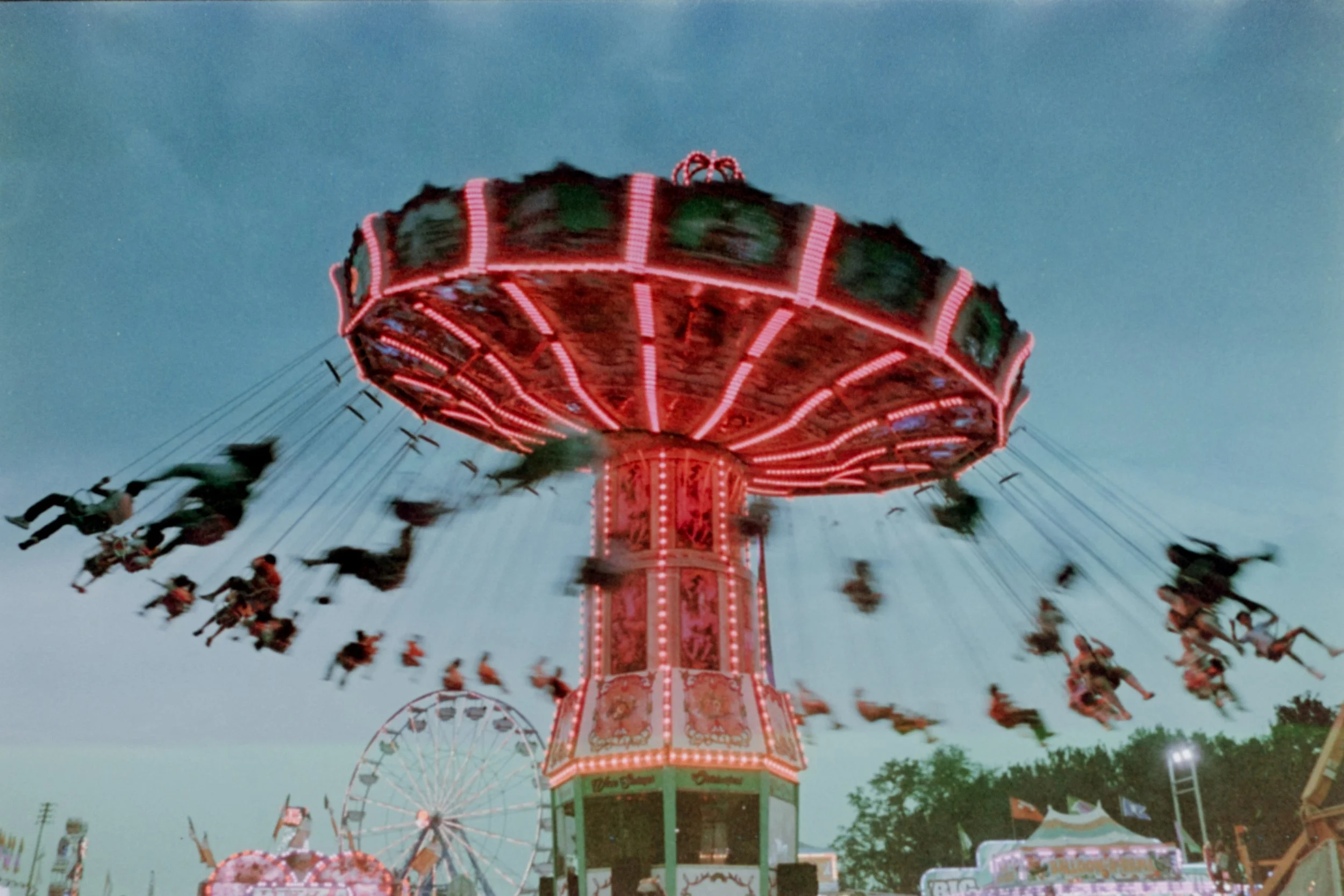 A brightly lit amusement park ride with swings spinning in the air, surrounded by other attractions and a ferris wheel, during twilight or early evening.