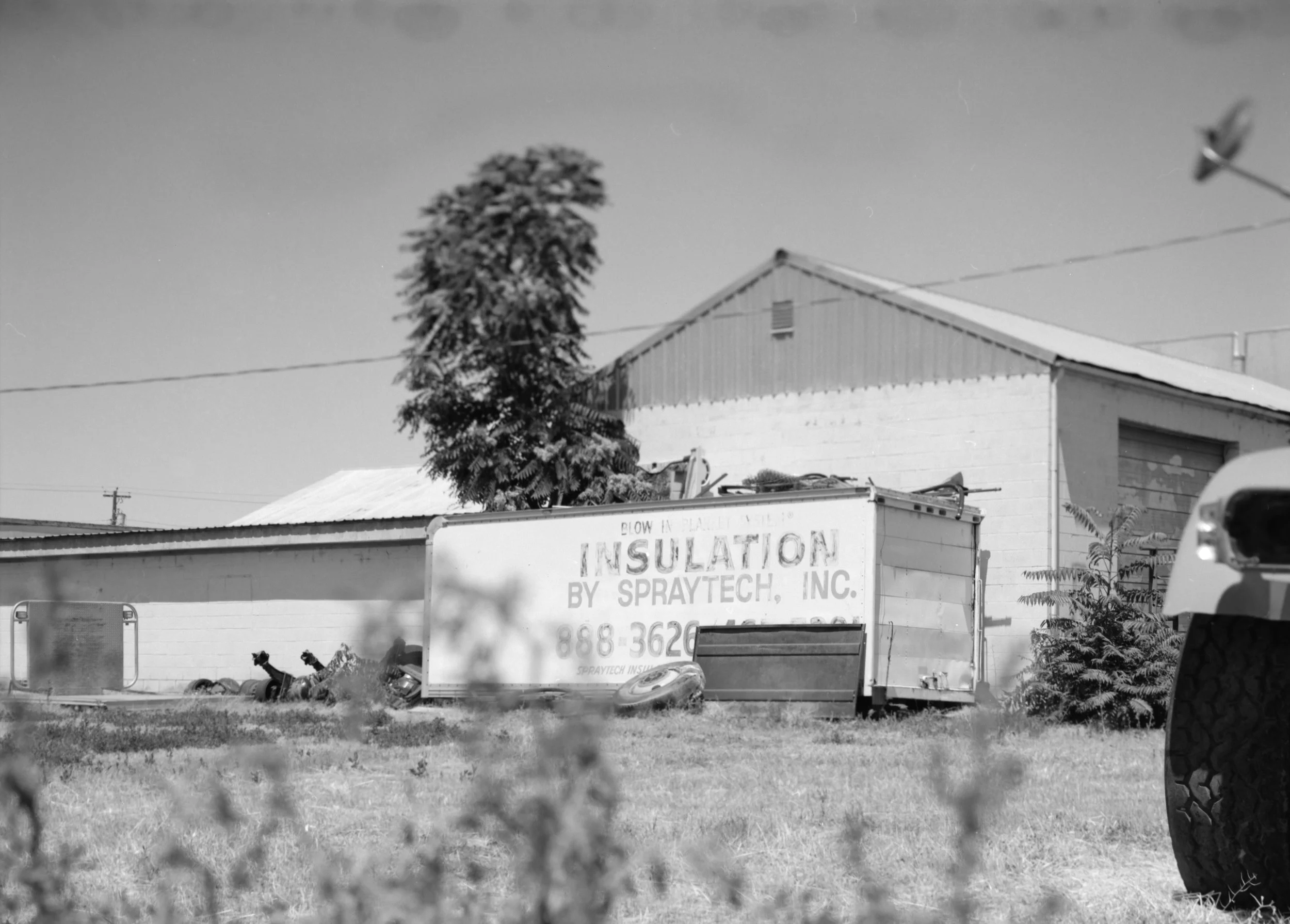 A black and white photo of a building with a sign that says 'Insulation by Spraytech, Inc.' and a phone number. There is a tree next to the building, and some items and a tire are on the ground in front. The foreground has some blurred plants, and a part of a vehicle side is visible on the right.