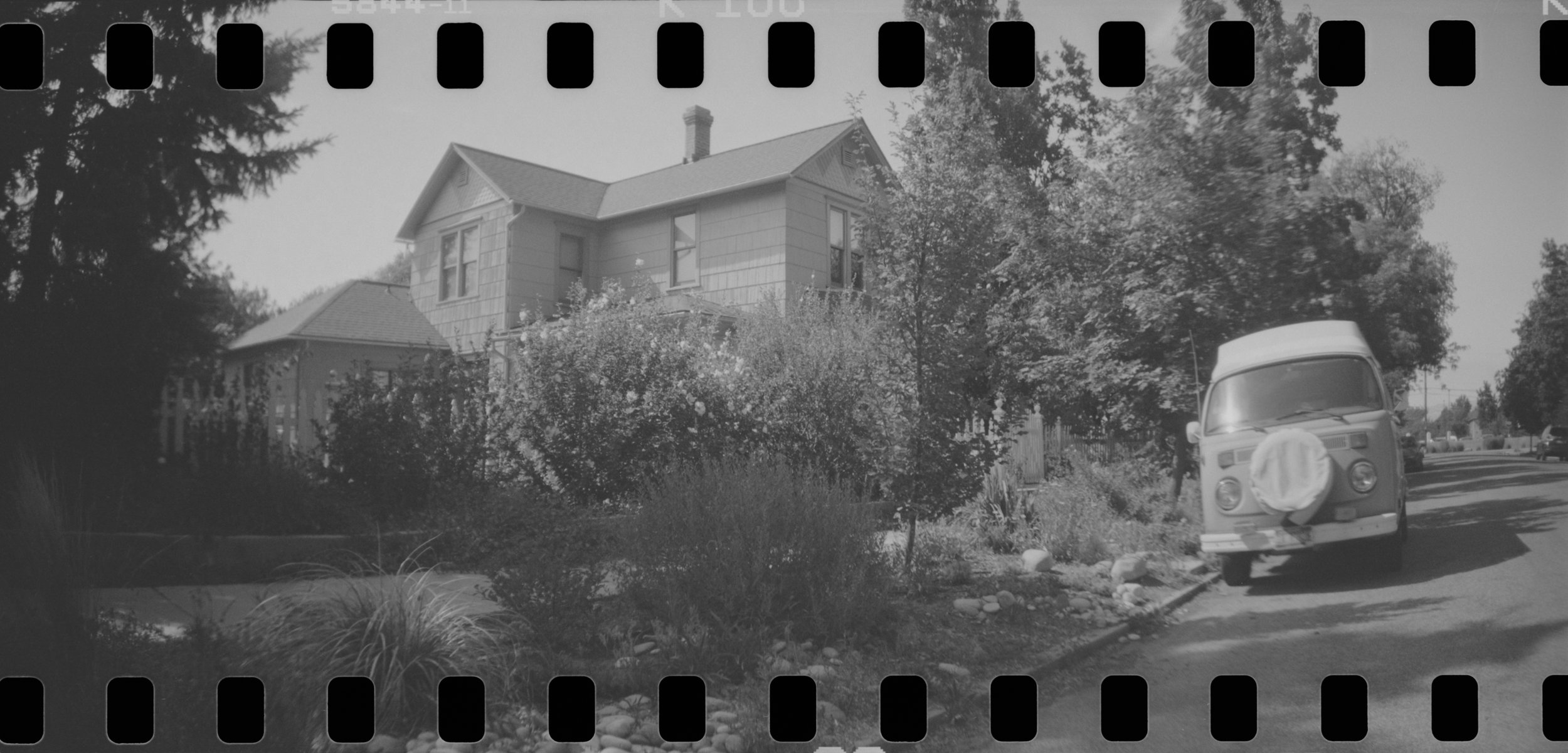 A black and white photograph of a residential street scene featuring a house with a pitched roof, shrubbery, a large tree, and a van with a covered spare tire parked on the road.