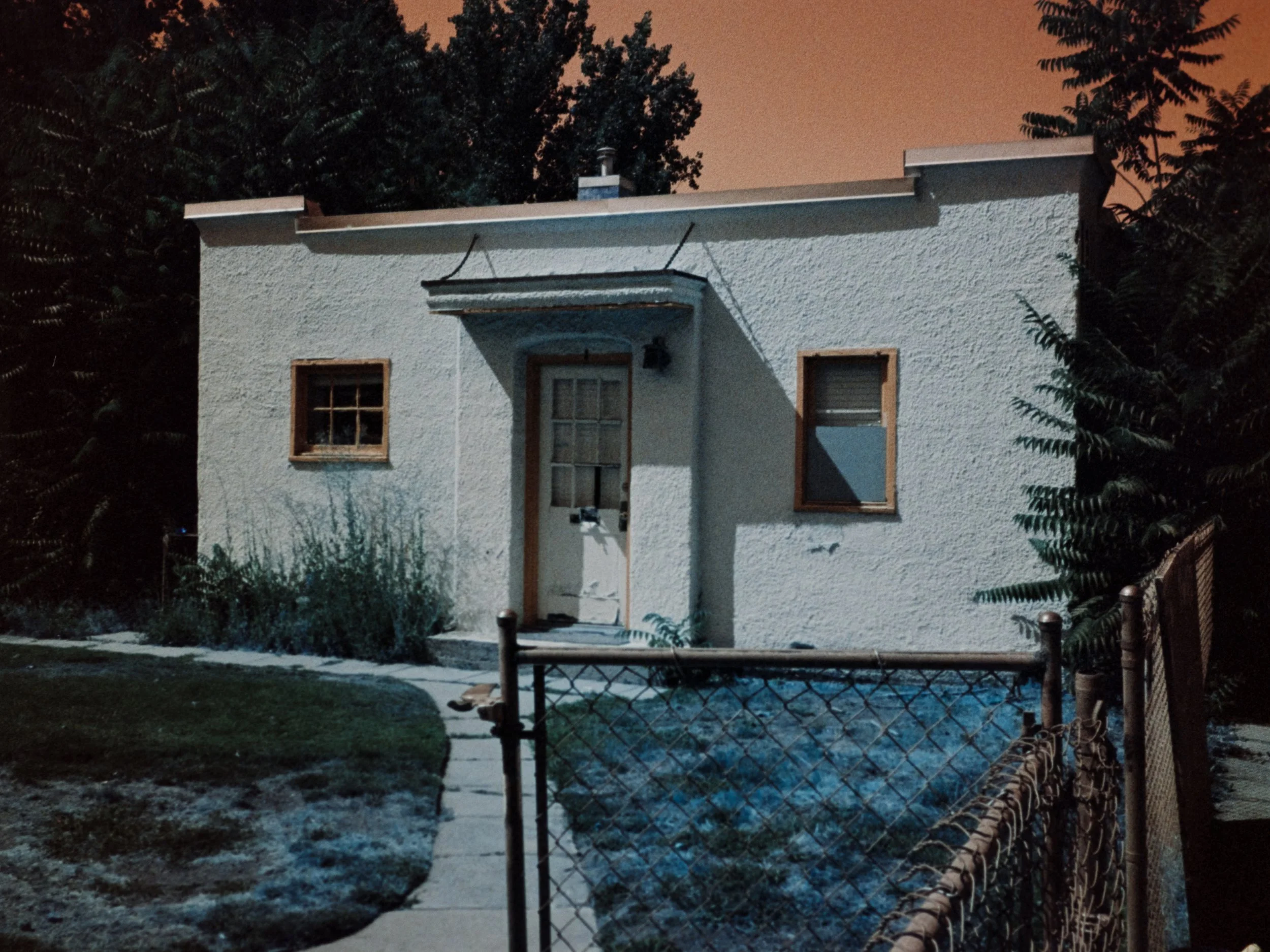 A small, white stucco house at night with two windows and a door, surrounded by trees and a chain-link fence.