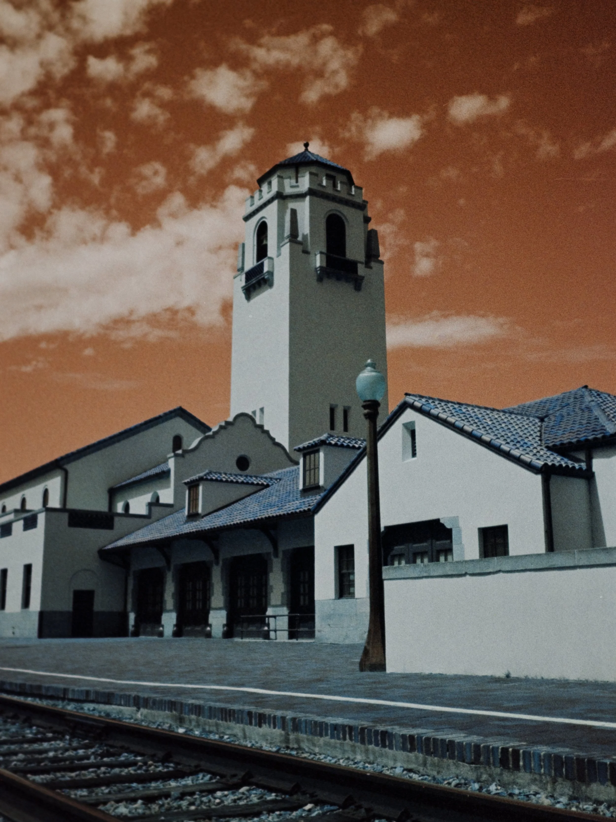 A tall, white clock tower with a dark blue roof stands behind a railway station platform with a streetlamp in the foreground. The sky is orange with scattered clouds.