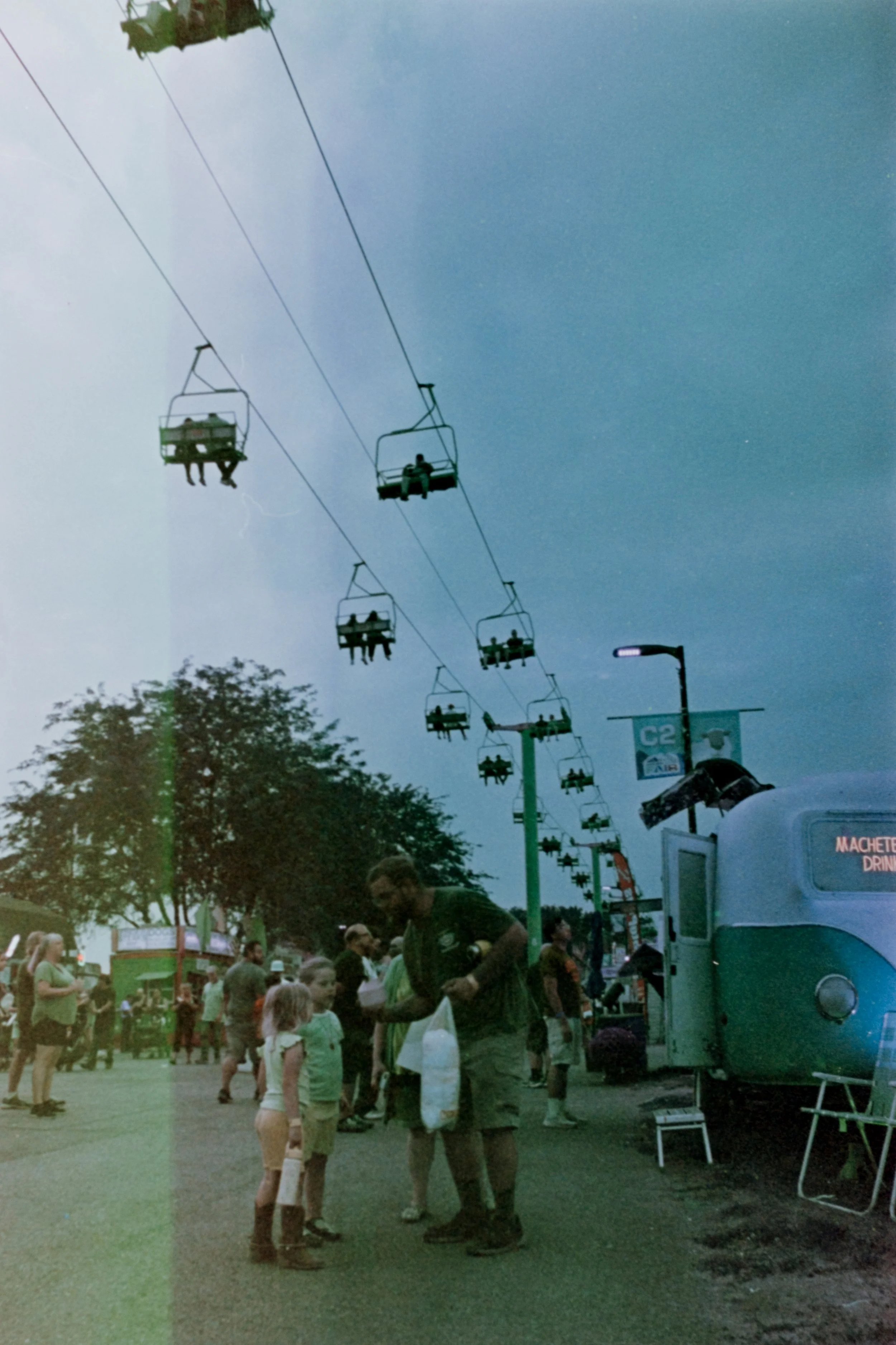 People at an outdoor fair or carnival, with a chairlift ride overhead and a food truck on the right side of the image.