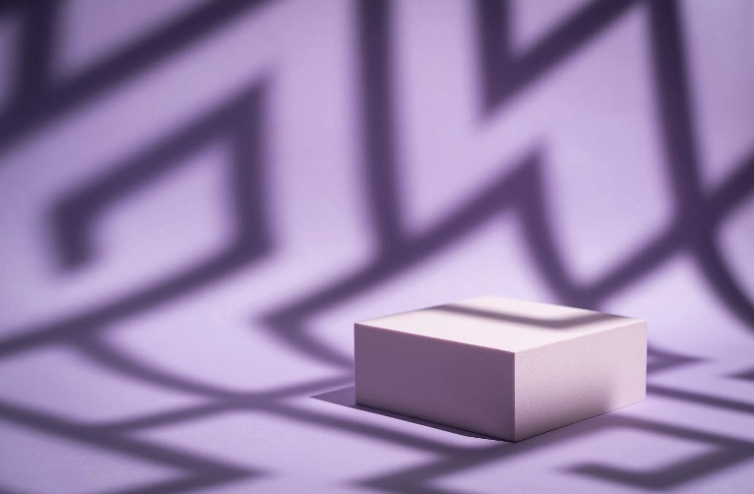 Caja blanca en una superficie con sombras geométricas formadas por una rejilla negra proyectadas en un fondo morado