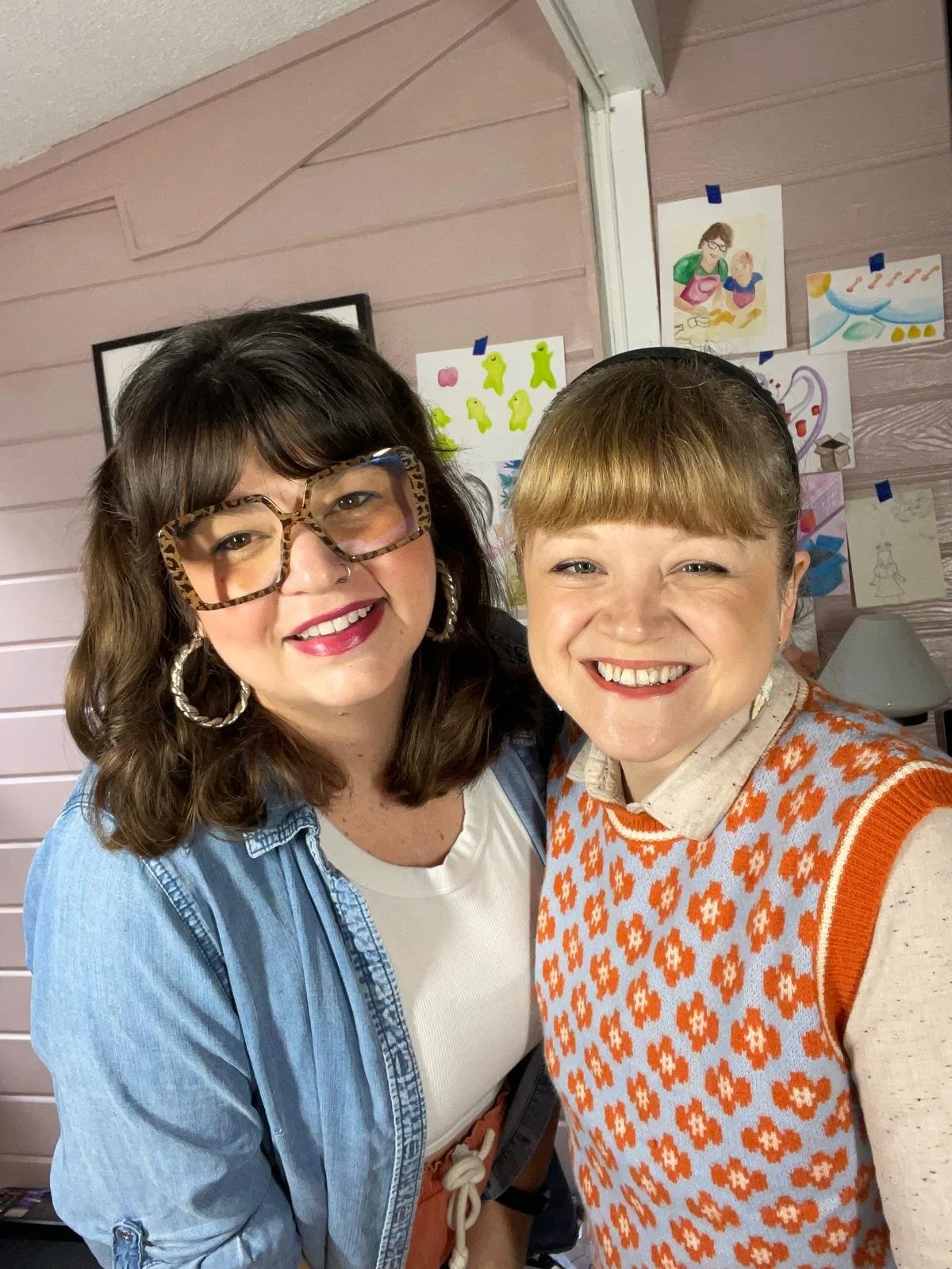 Two women, Becky and Mandi Meadows, smiling and posing for a photo in front of colorful children's drawings taped to a pink wooden wall.