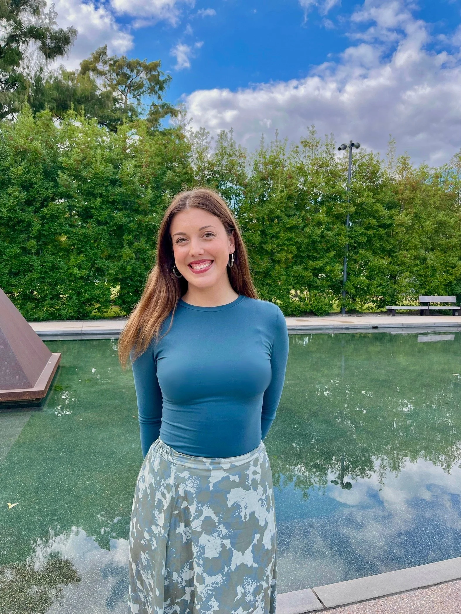 A smiling young woman with long brown hair, wearing a blue long-sleeved top and a patterned skirt, standing outdoors near a reflective pool with lush green trees in the background under a partly cloudy sky.