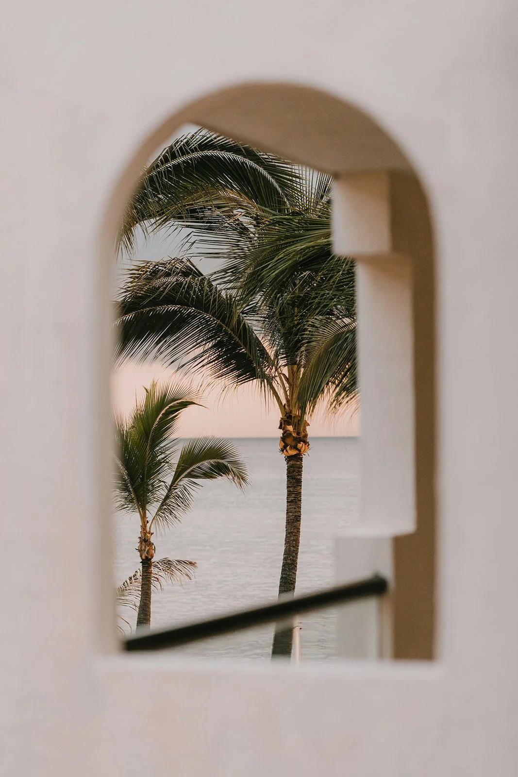 View through an arched wall opening of palm trees near a calm seaside at sunset.