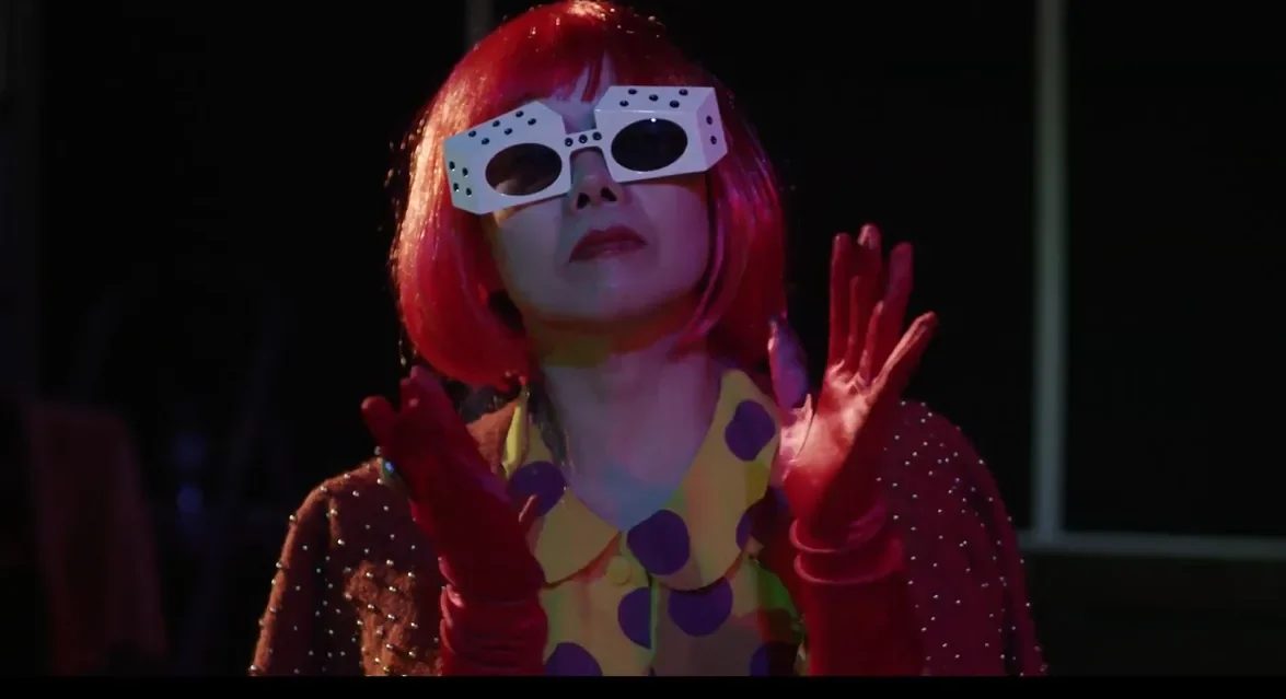 A red-haired performer wearing oversized white sunglasses, a polka-dot dress and red gloves in a film scene.
