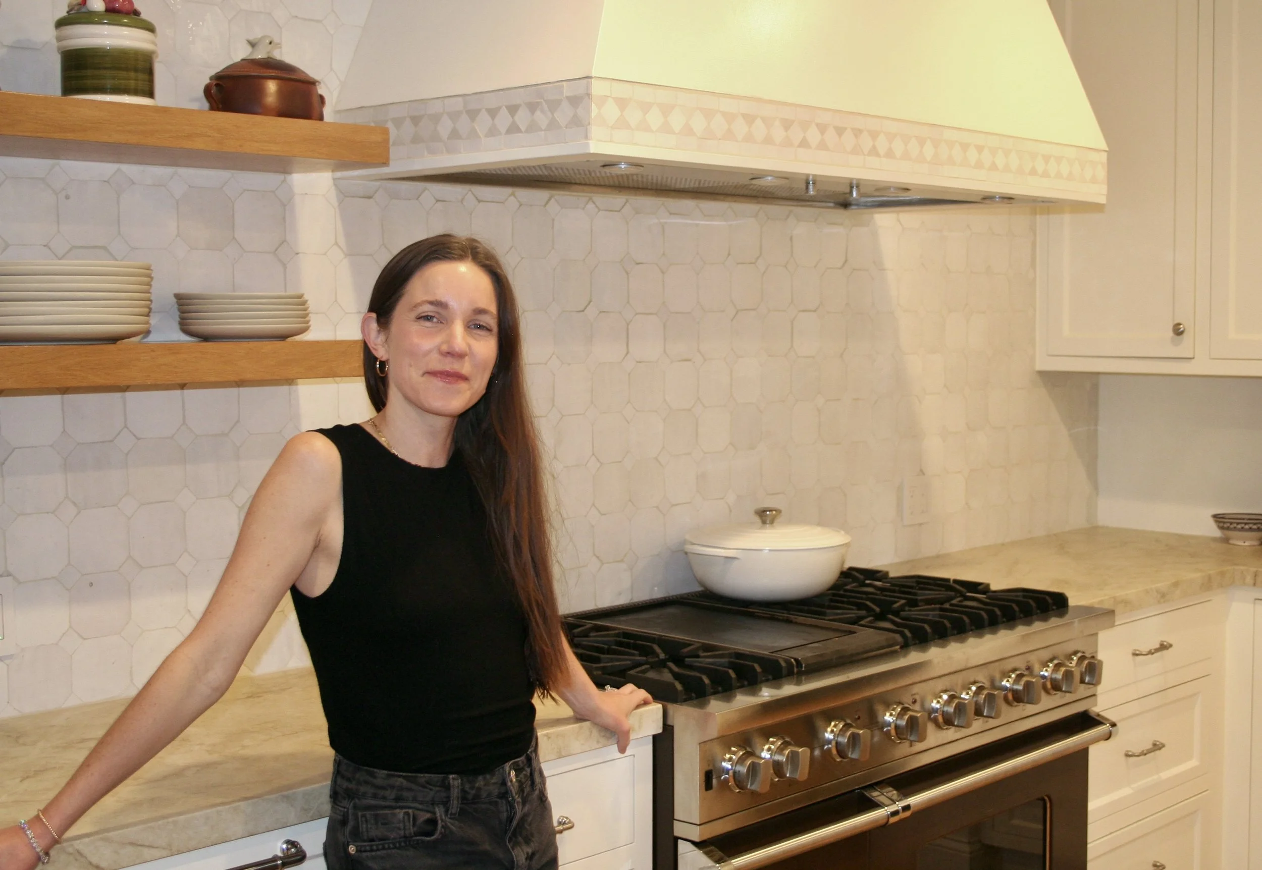 A woman with long brown hair wearing a black sleeveless top stands in a kitchen with cream-colored cabinets, a stovetop, and open wooden shelves with dishes and decorative items.