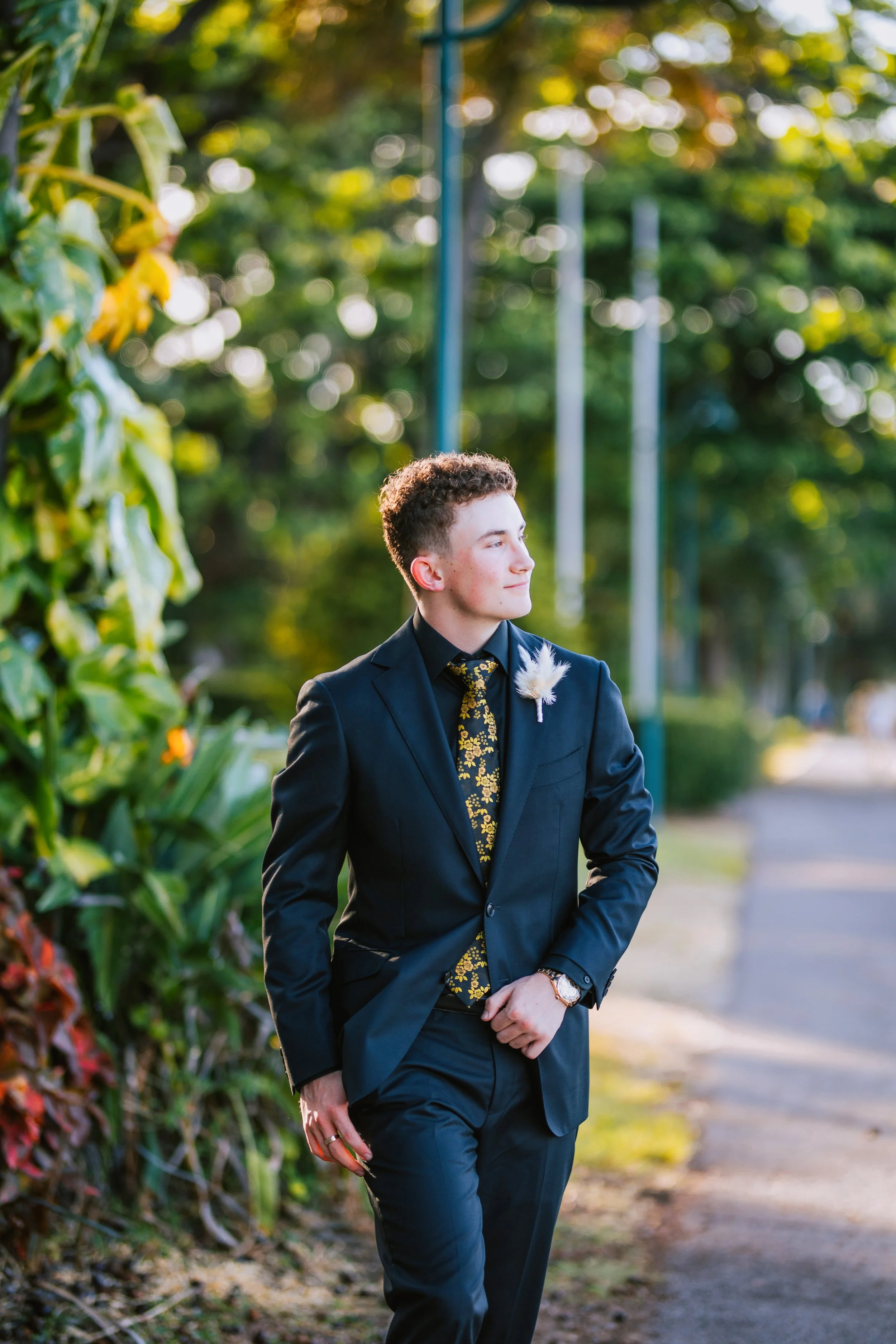 A young man in a black suit with a floral tie and a feather boutonniere stands outdoors on a sidewalk, with green trees and streetlamps in the background during sunset.