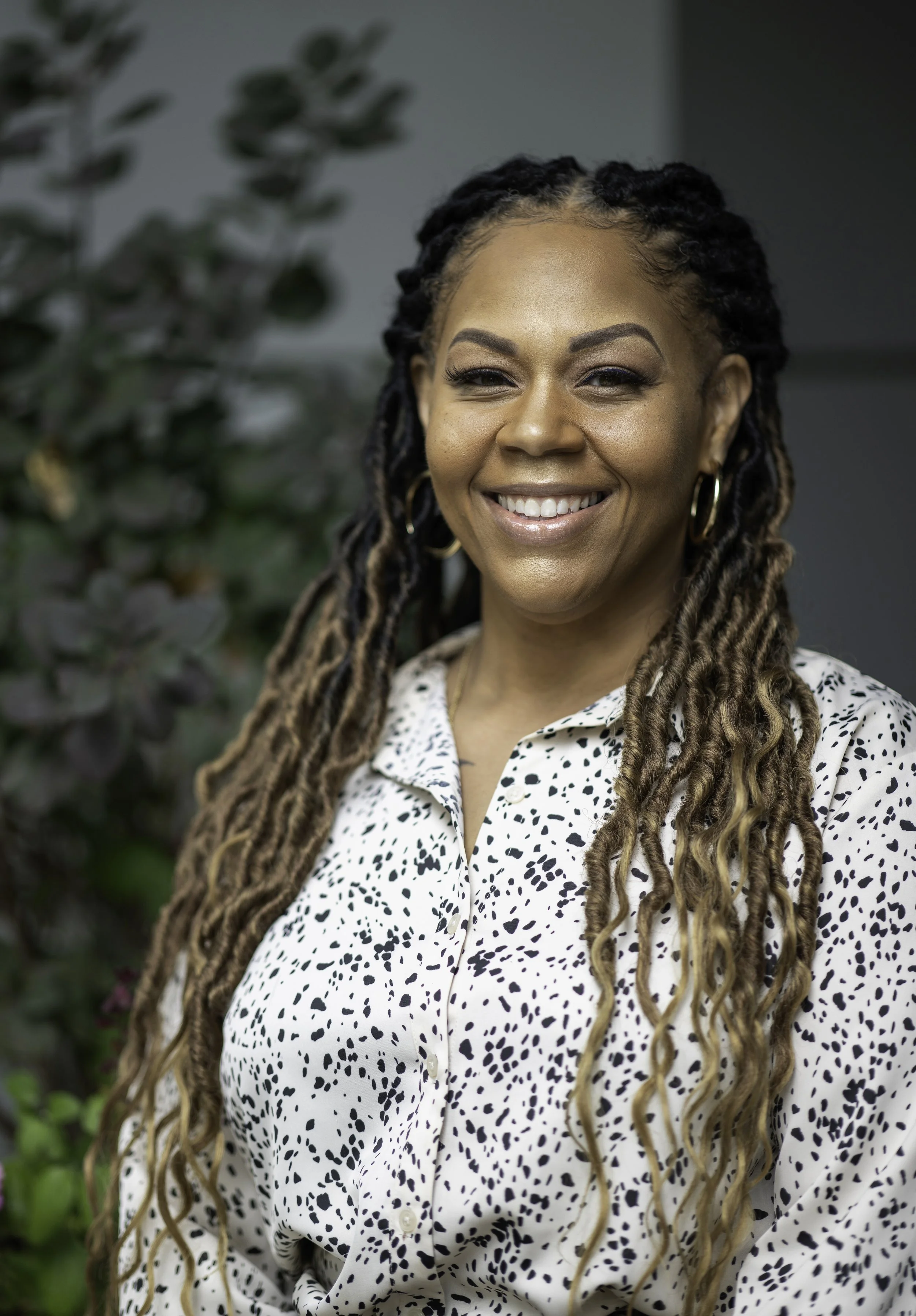 A woman with long, curly dreadlocks smiling at the camera, wearing a white blouse with black irregular spots, gold hoop earrings, and standing in front of leafy green foliage.