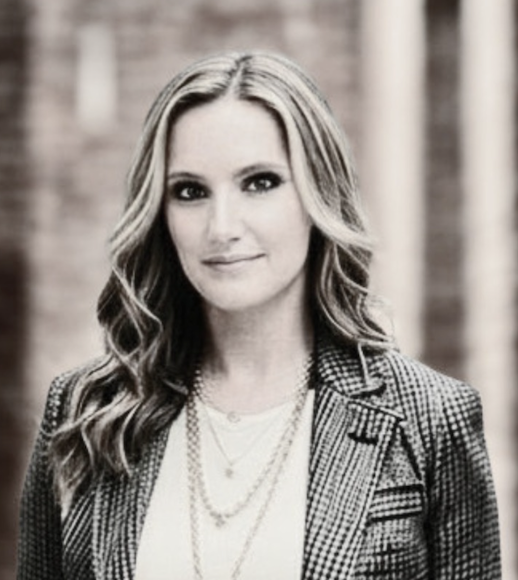 Black and white portrait of a woman with wavy hair, wearing a checked blazer and layered necklaces.