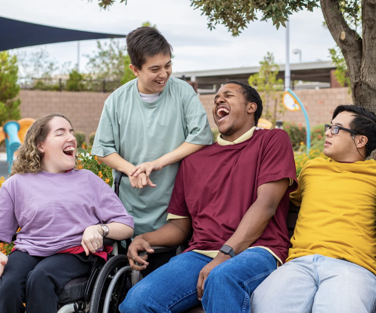 Four young people outdoors, laughing and enjoying each other's company in a park or playground, with a tree and playground equipment in the background.