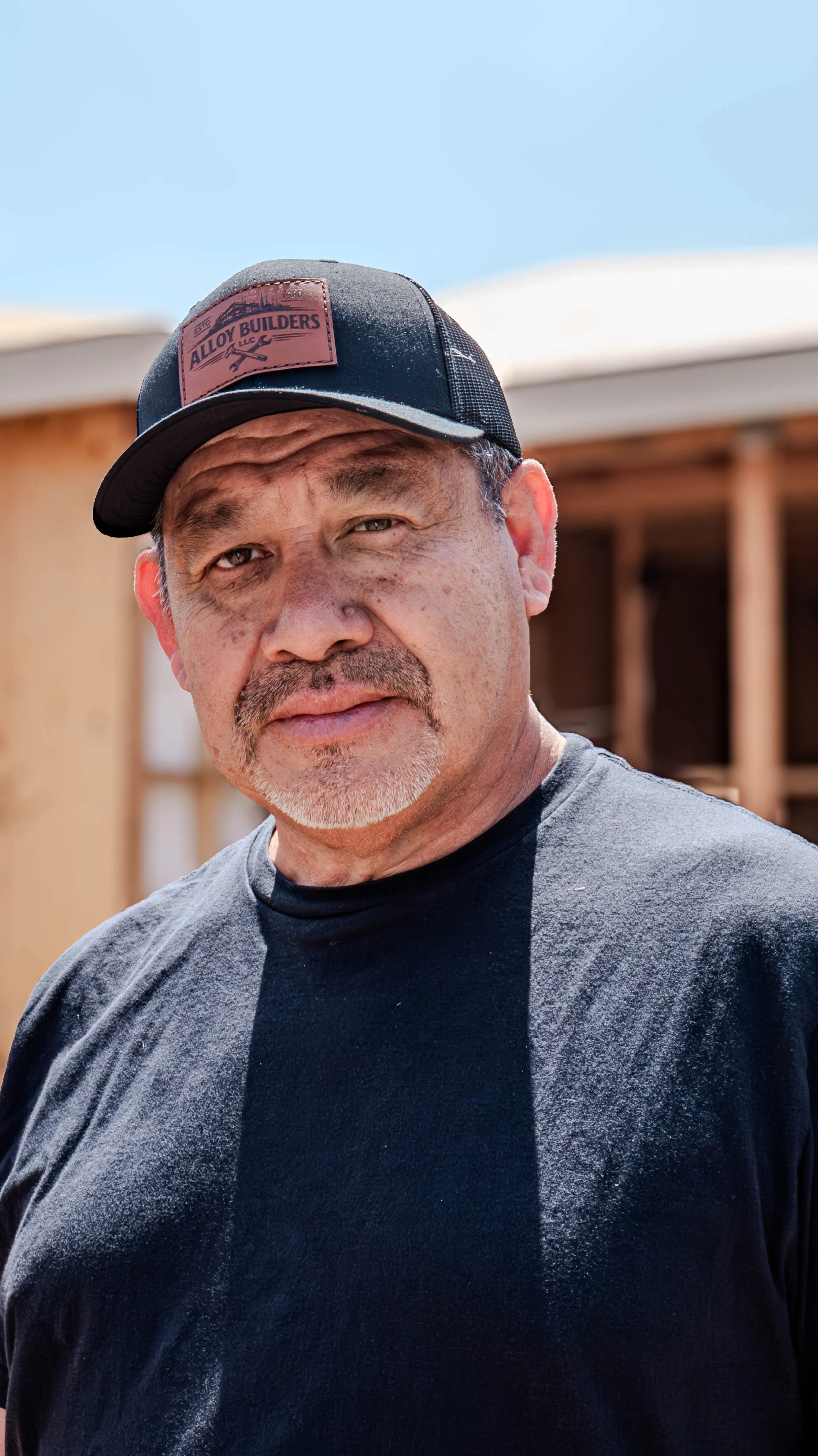 A middle-aged man with light brown eyes, a gray goatee, and salt-and-pepper hair stands outdoors, wearing a black T-shirt and a black trucker hat with a brown patch that reads 'ALLOY BUILDERS LLC' and shows a wrench and screwdriver crossed.