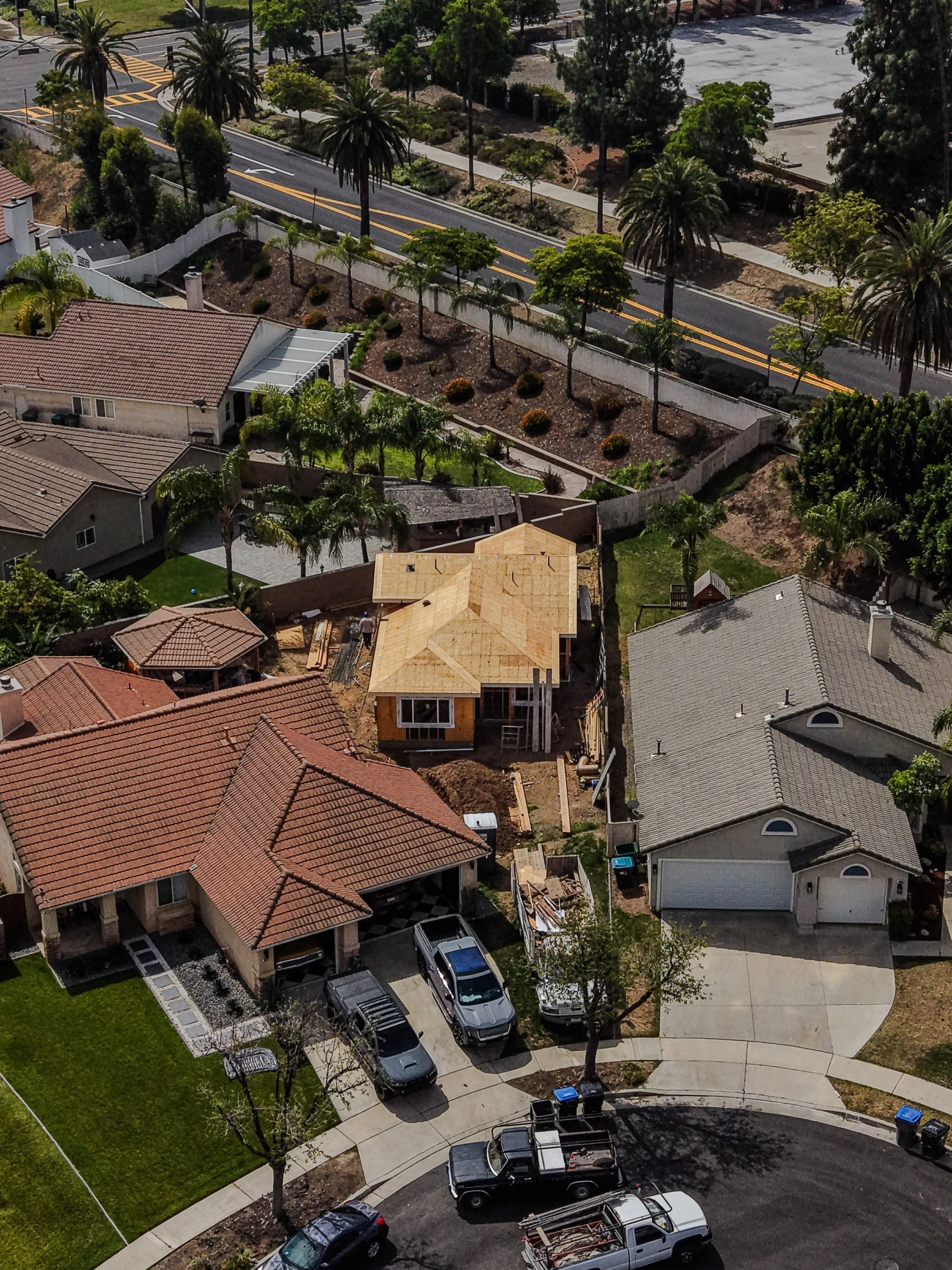 A neighborhood with houses, trees, and parked cars. A house under construction is visible with a wooden roof framework. There are paved sidewalks, a street with trees, and a parking lot in the background.