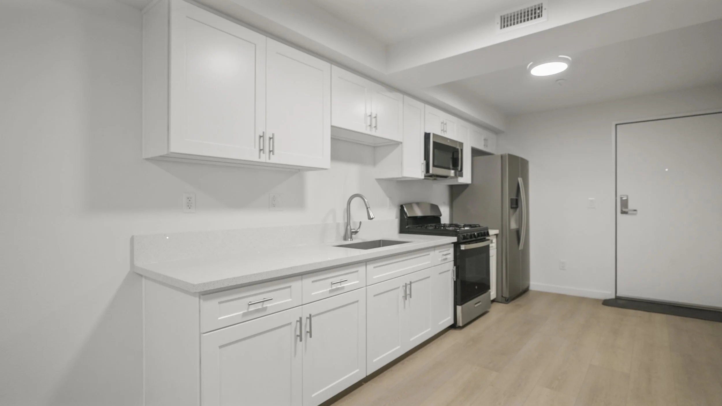 Modern kitchen with white cabinets, a gray refrigerator, stovetop, oven, microwave, and a stainless steel sink with a faucet, light wood flooring, and a white door.