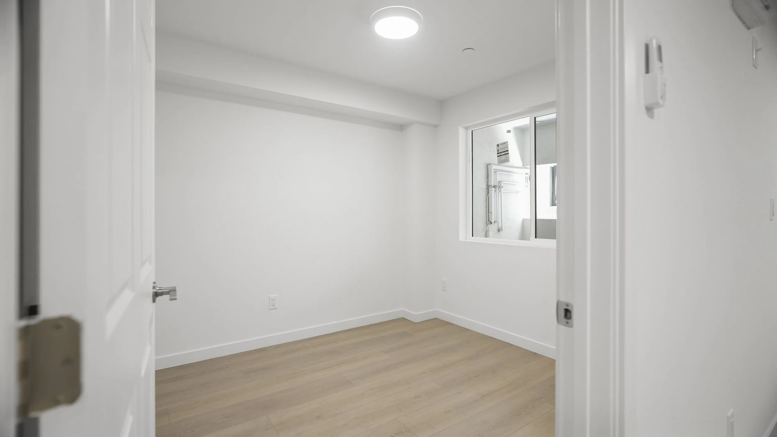 Empty white room with a window and light wood flooring, viewed from an open door.