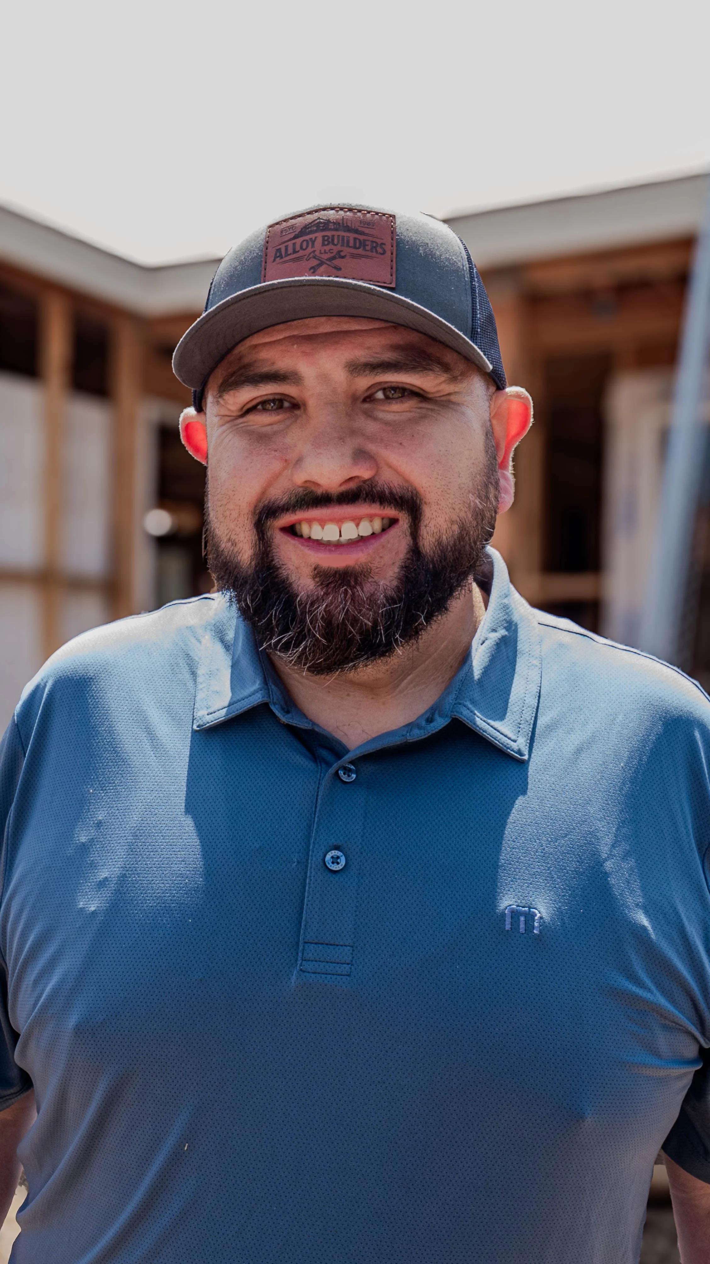 A smiling man with a beard wearing a blue polo shirt and a gray and blue baseball cap outdoors.