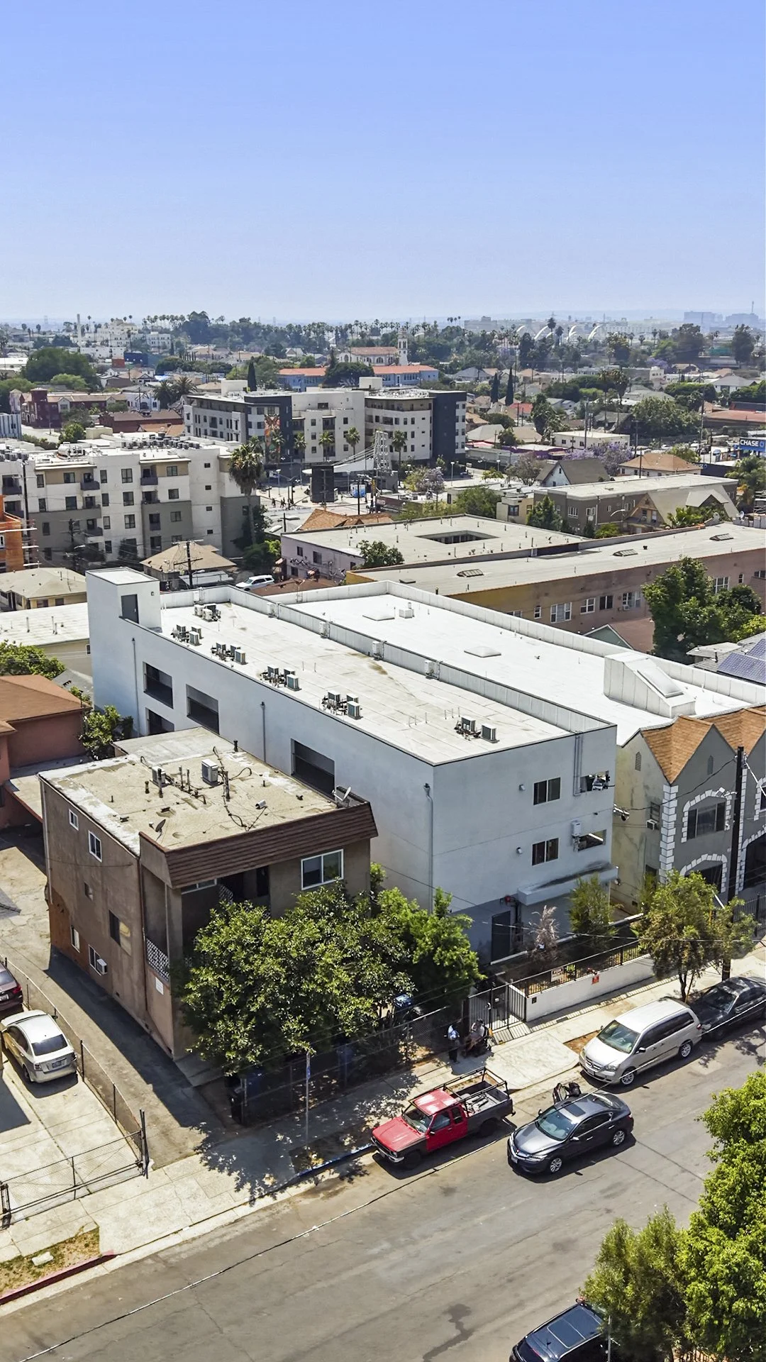 Aerial view of a cityscape showing multiple buildings, trees, streets with parked cars, and a distant horizon with more buildings and trees under a clear blue sky.