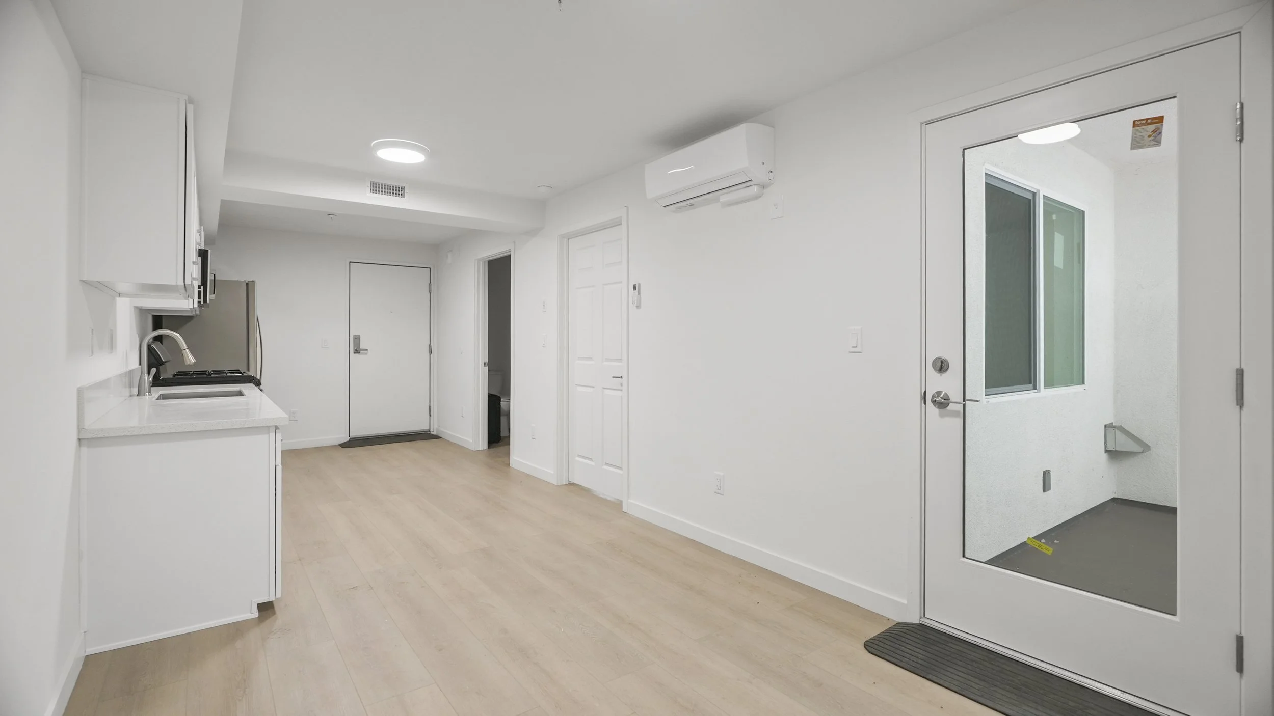 Interior of a small, modern apartment or studio with white walls, light wood flooring, a kitchenette on the left, an air conditioning unit on the wall, and a glass door leading outside.