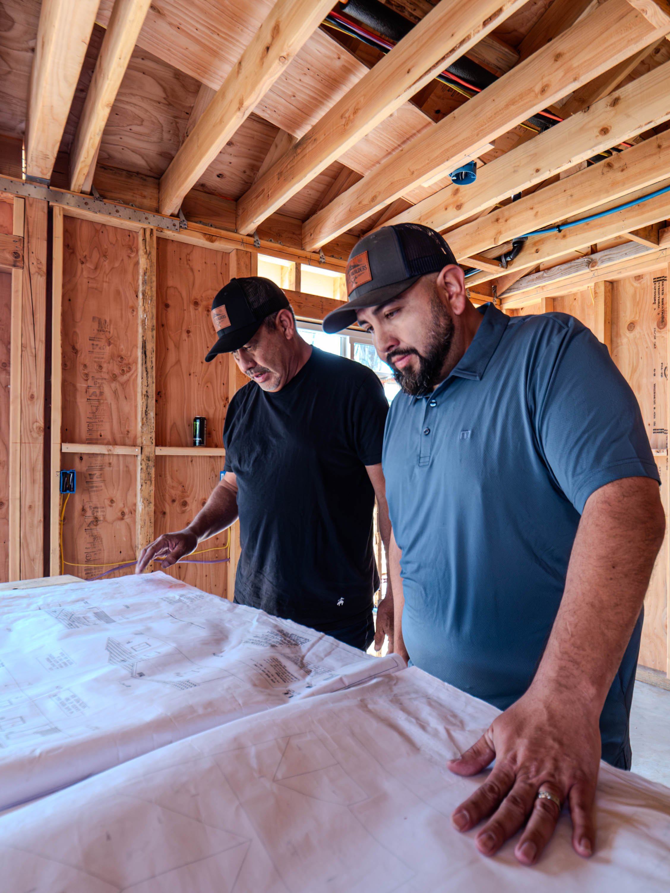 Two men in baseball caps examining construction blueprints on a table inside a wooden framed building under construction.