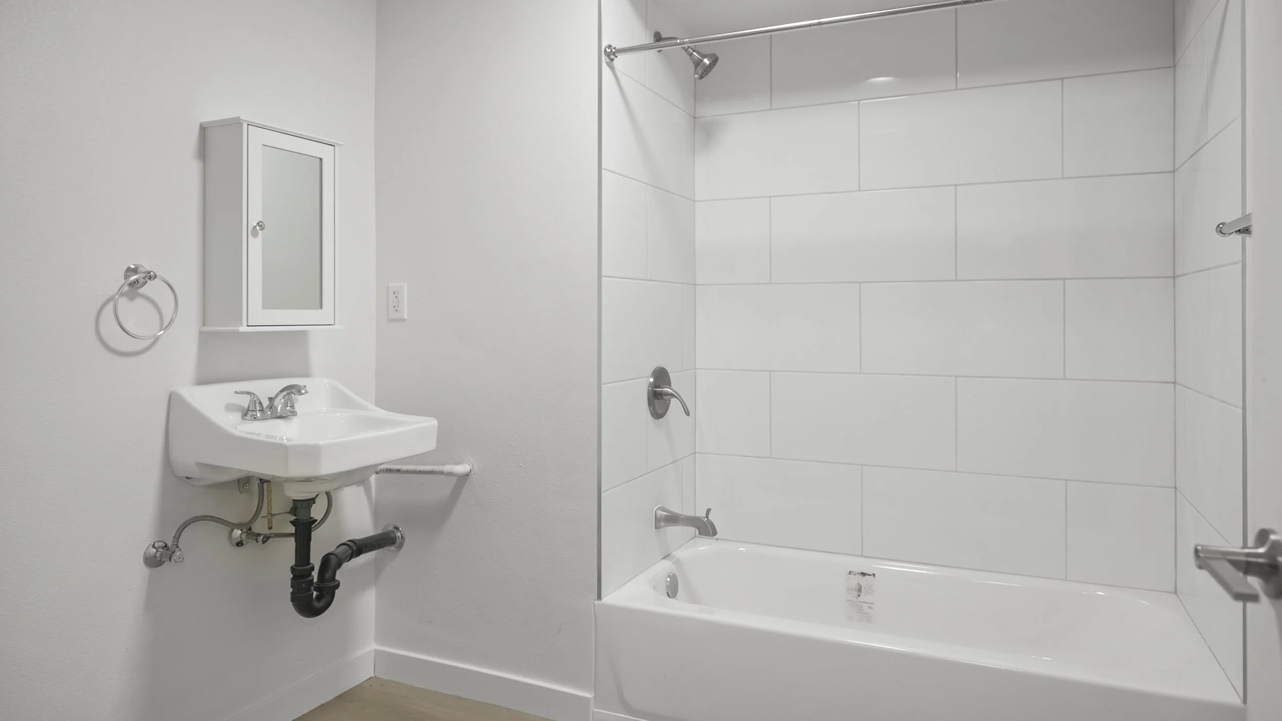 Bathroom with a small wall-mounted sink, a mirror cabinet, a towel ring, and a bathtub with a showerhead and a grab bar, white tiled walls and light-colored flooring.