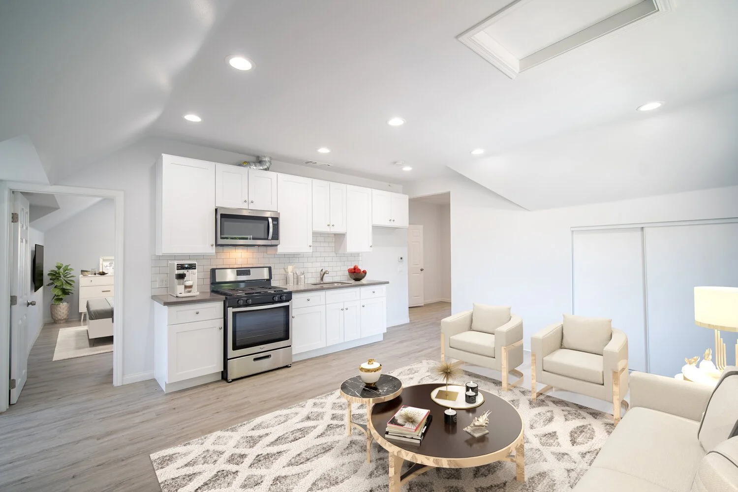 Bright living room with white walls, ceiling, and kitchen cabinets; gray wooden floor; black and white area rug; cream-colored armchairs and sofa; black coffee table with decorative items; kitchen area with stove, microwave, coffee maker, and bowl of red apples.