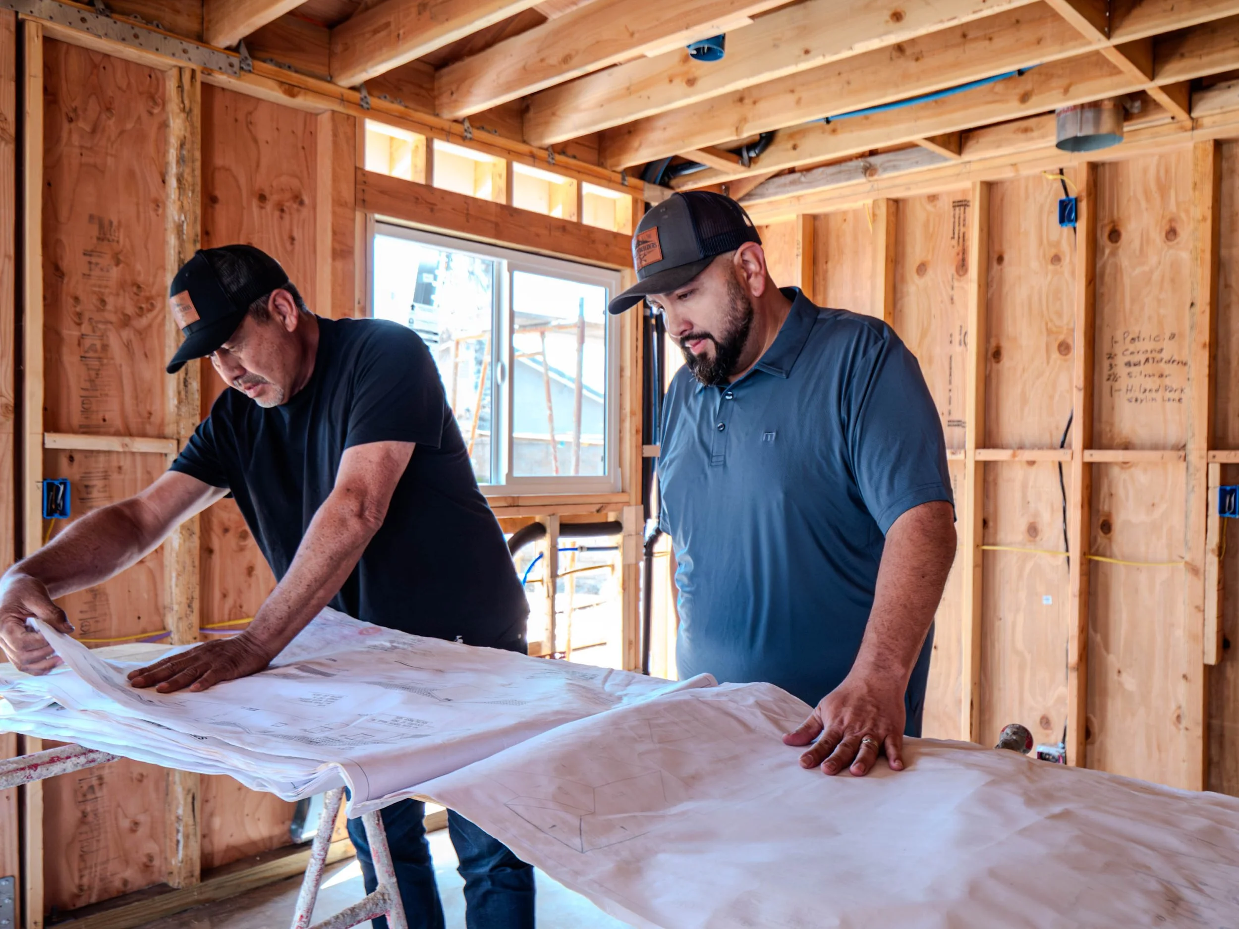 Two men wearing black and gray caps and dark shirts look at a blueprint or plan on a table inside a building under construction with exposed wooden framing and a window in the background.