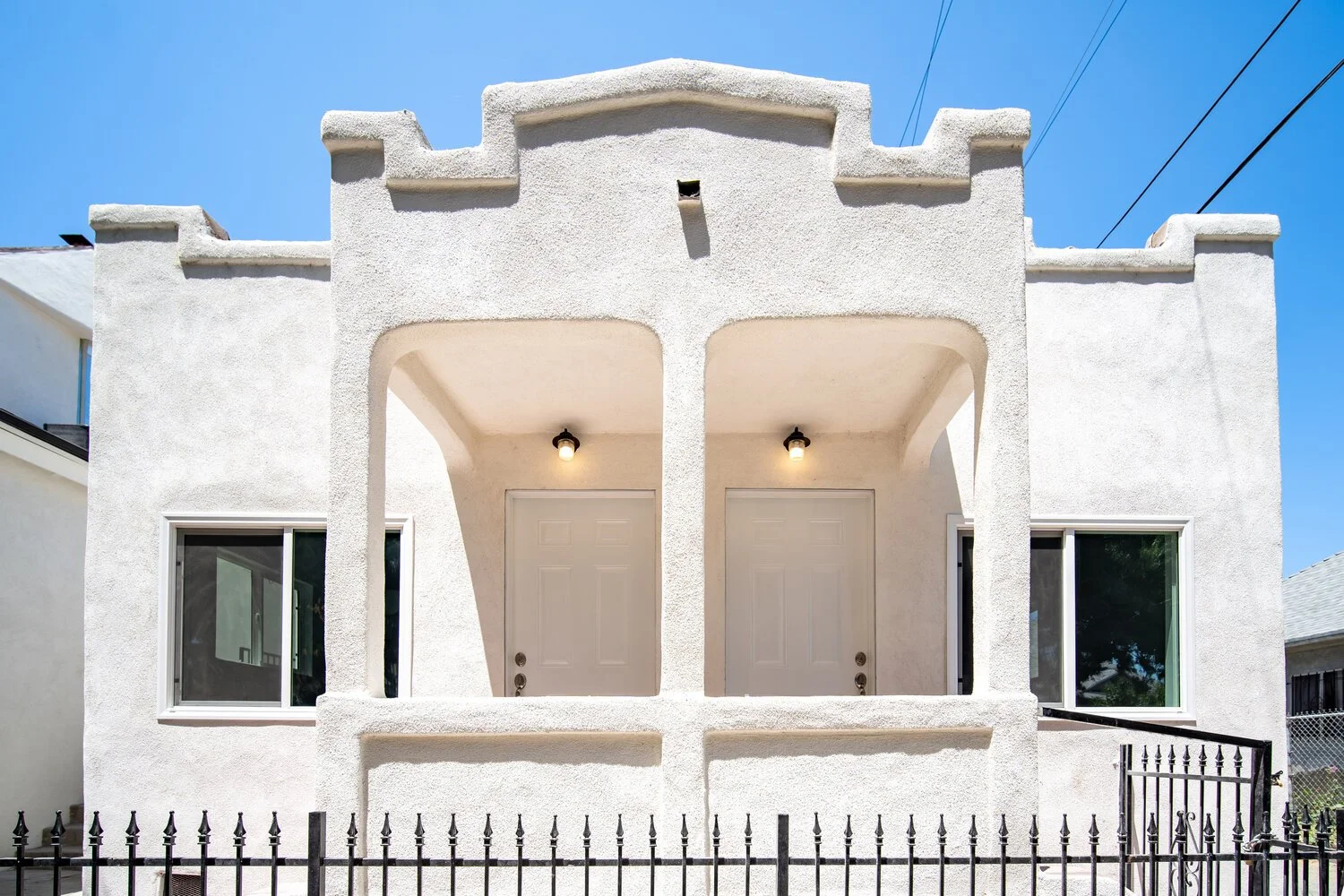White two-story building with arched entryways, two front doors, and two windows, surrounded by a black iron fence, under a clear blue sky.