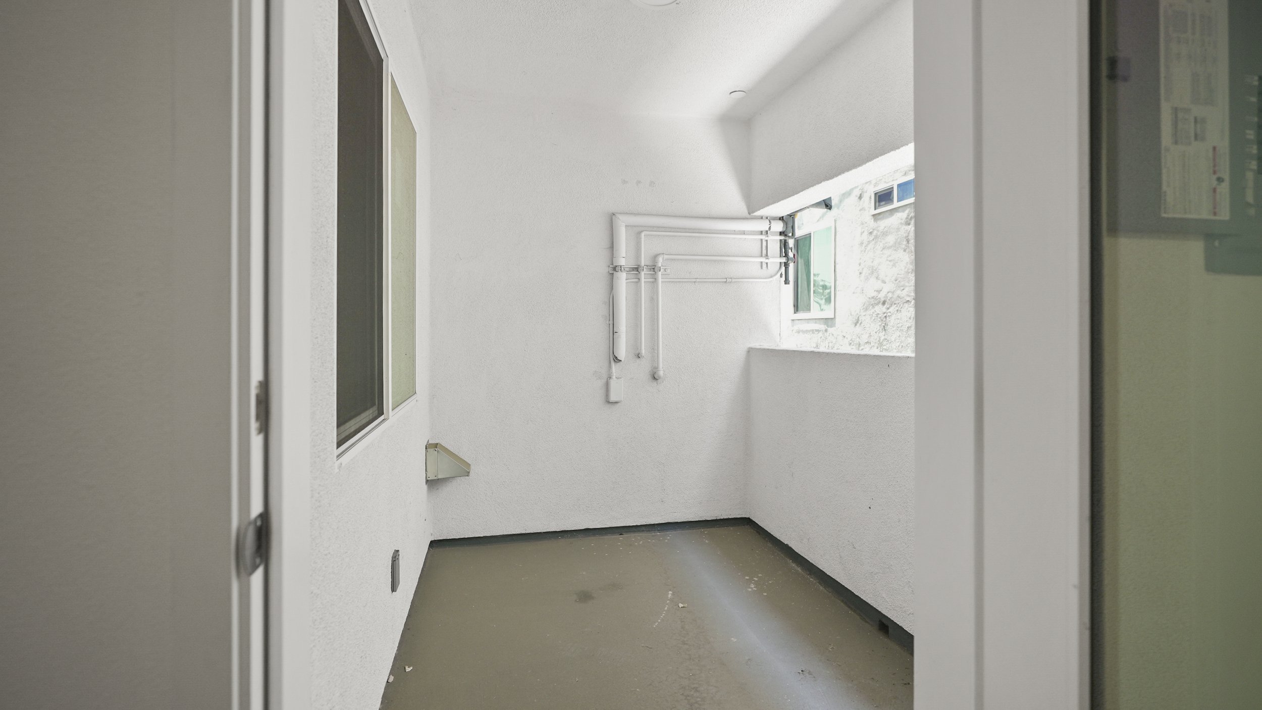Empty outdoor balcony with white walls, two windows, pipe fixtures, and a concrete floor with some debris.