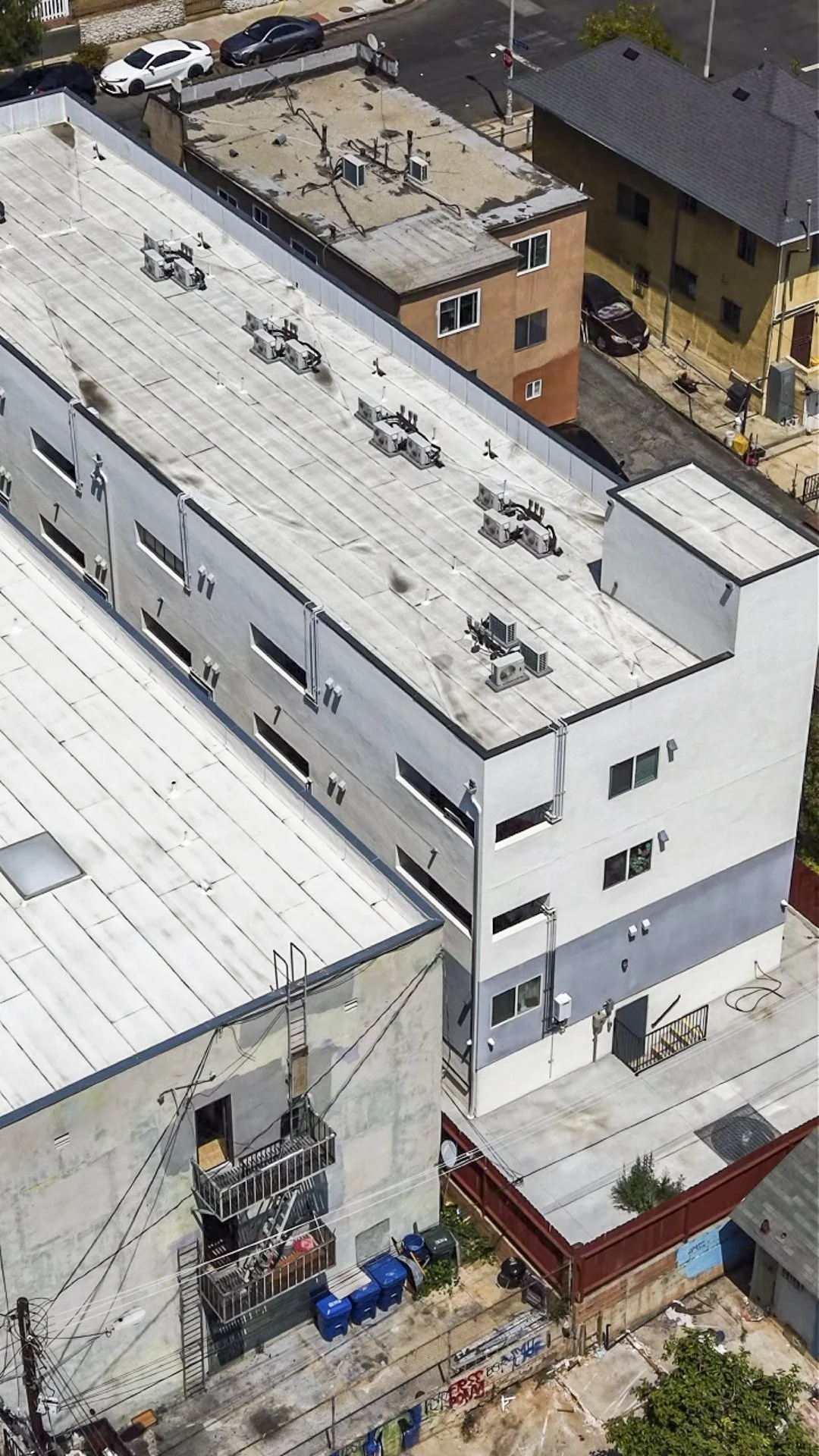 Aerial view of city buildings including a large white commercial or apartment building with vents and air conditioning units on the roof, nearby smaller residential buildings, parked cars, and a street with graffiti.