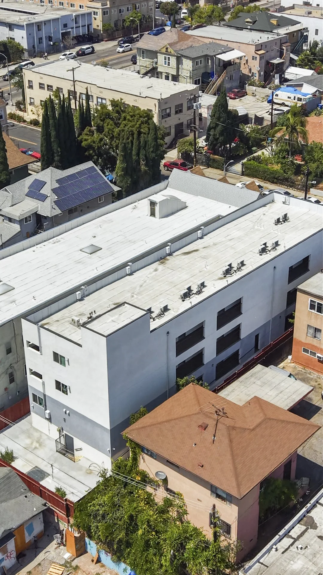 Aerial view of a city block with various residential buildings, including an apartment complex with a white flat roof, a house with solar panels, and other homes with differing Roof styles and colors.