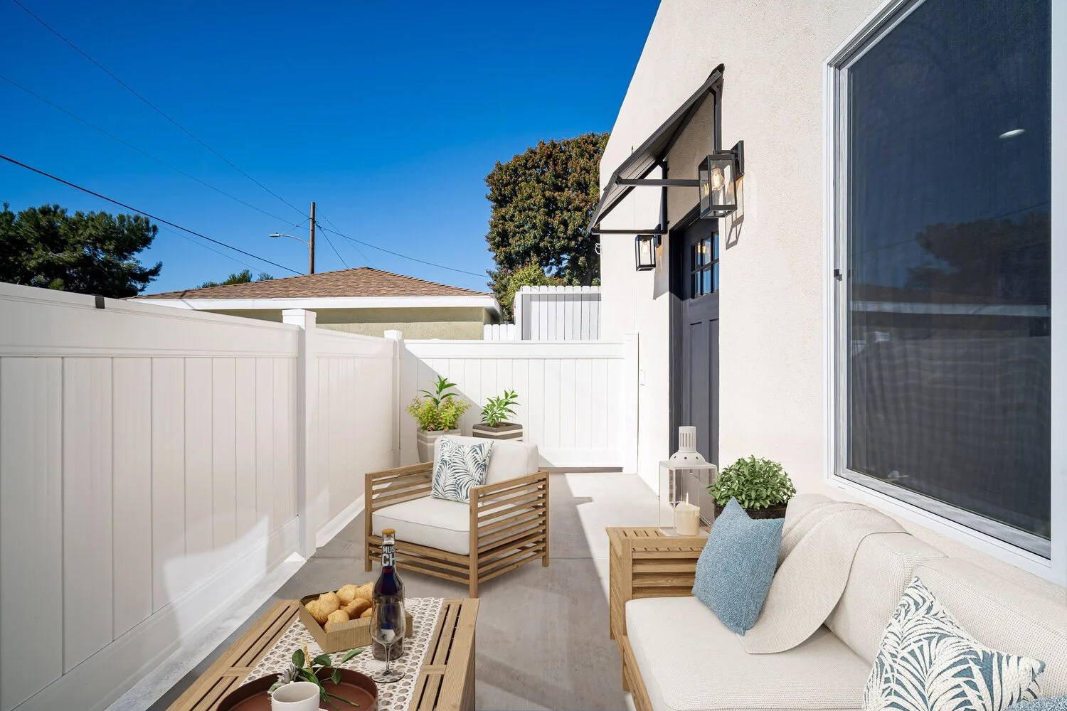 Decorative outdoor patio area with wooden furniture, potted plants, lantern, and a bottle of wine with snacks, enclosed by white fence under clear blue sky.