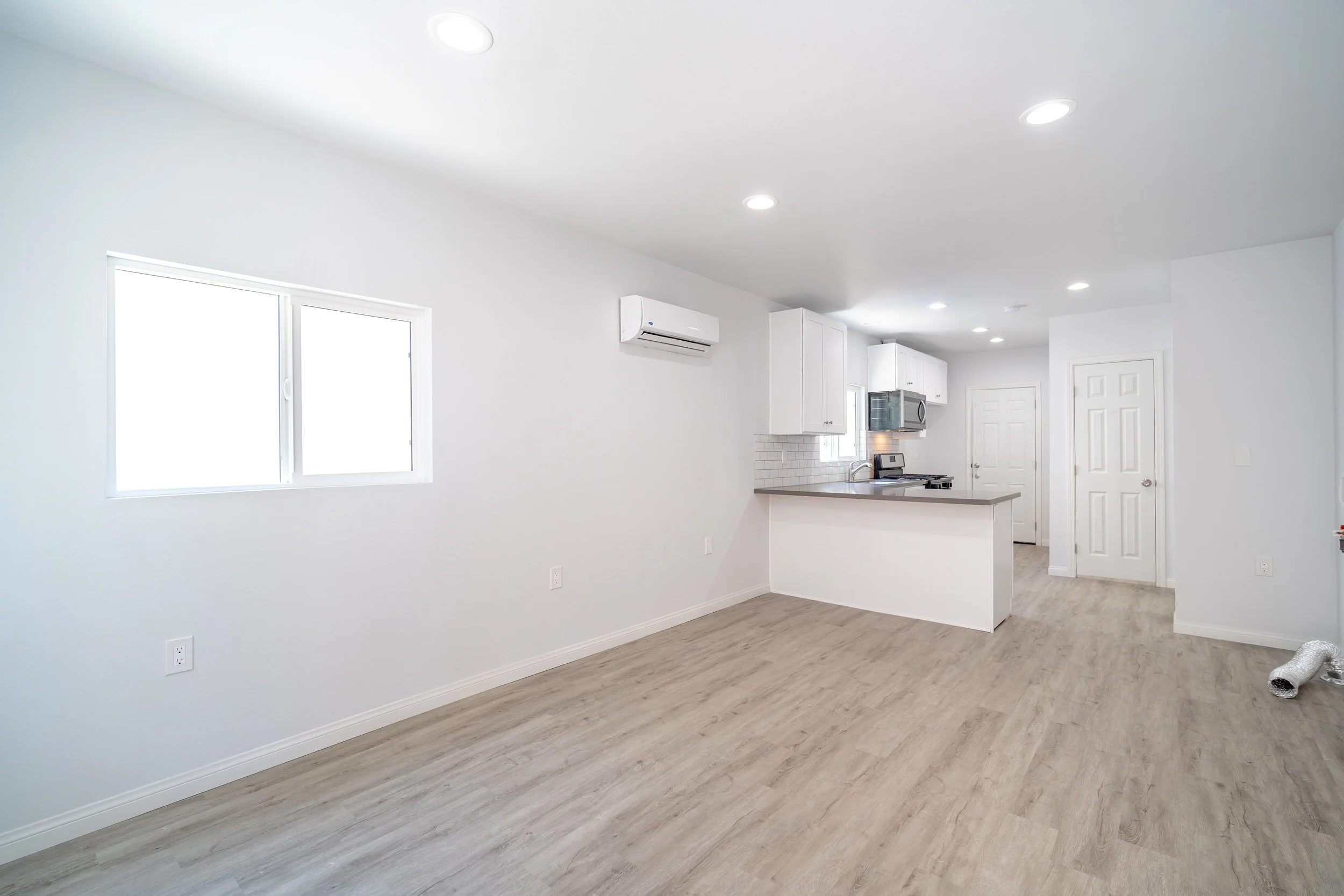 Empty kitchen with white walls, ceiling, and cabinets, a window, wood flooring, and ceiling lights, with a duct in the corner.