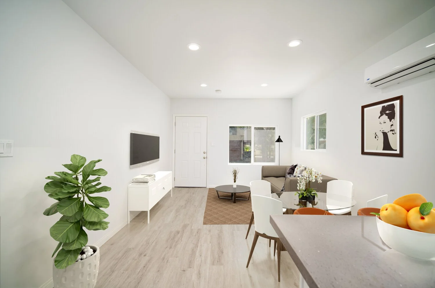 Bright living room with white walls, wooden flooring, TV on wall, beige sofa, round coffee table, dining area with white chairs, fruit bowl, potted plant, wall art, large windows, air conditioner.