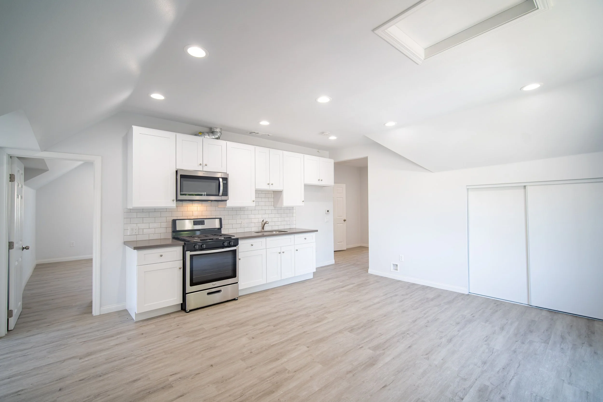 Empty modern kitchen with white cabinets, stainless steel appliances, and light wood flooring, with bright ceiling lights.