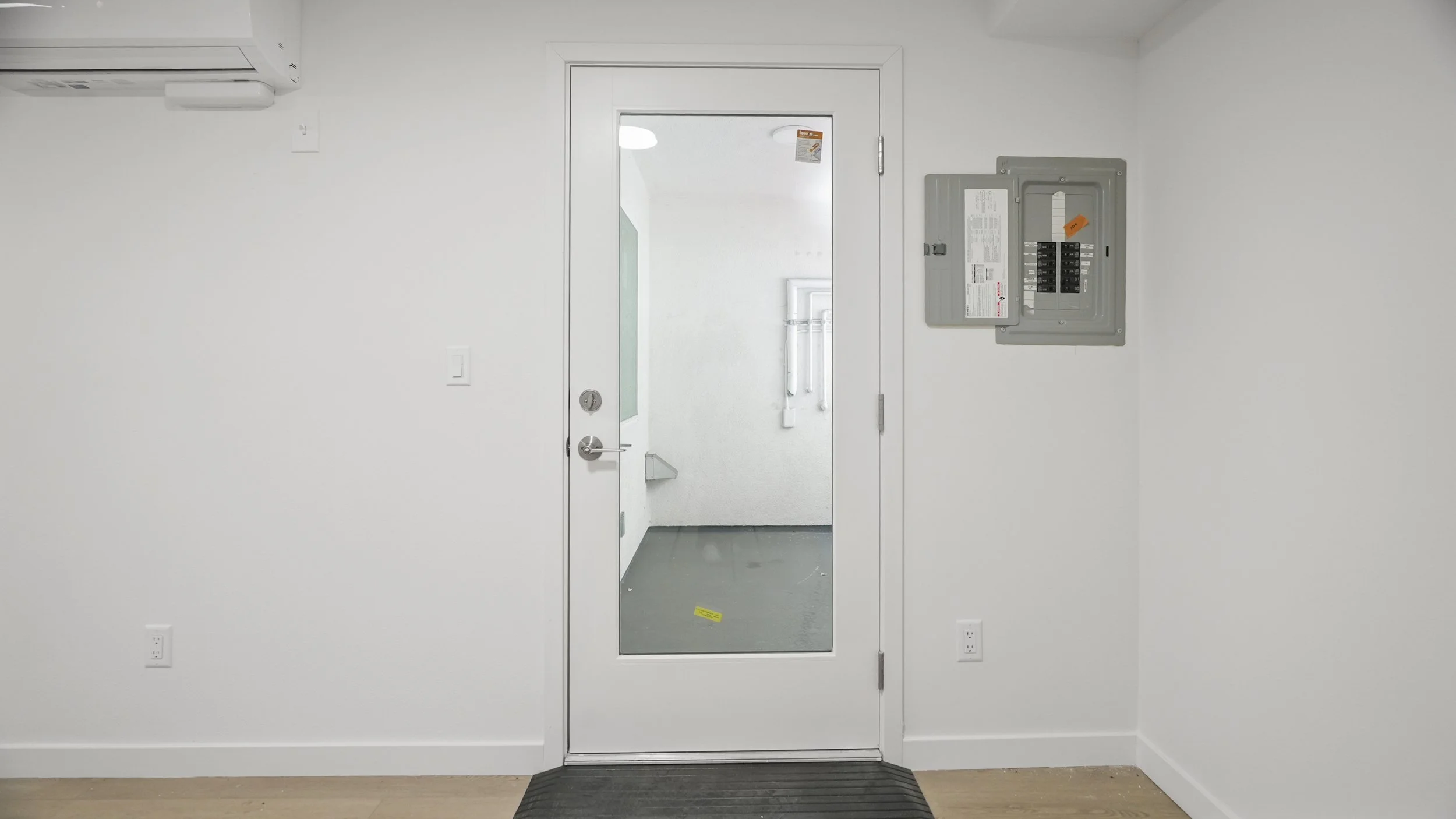 White wall with a glass door, electrical panel, air conditioning unit, and a gray mat on the floor.