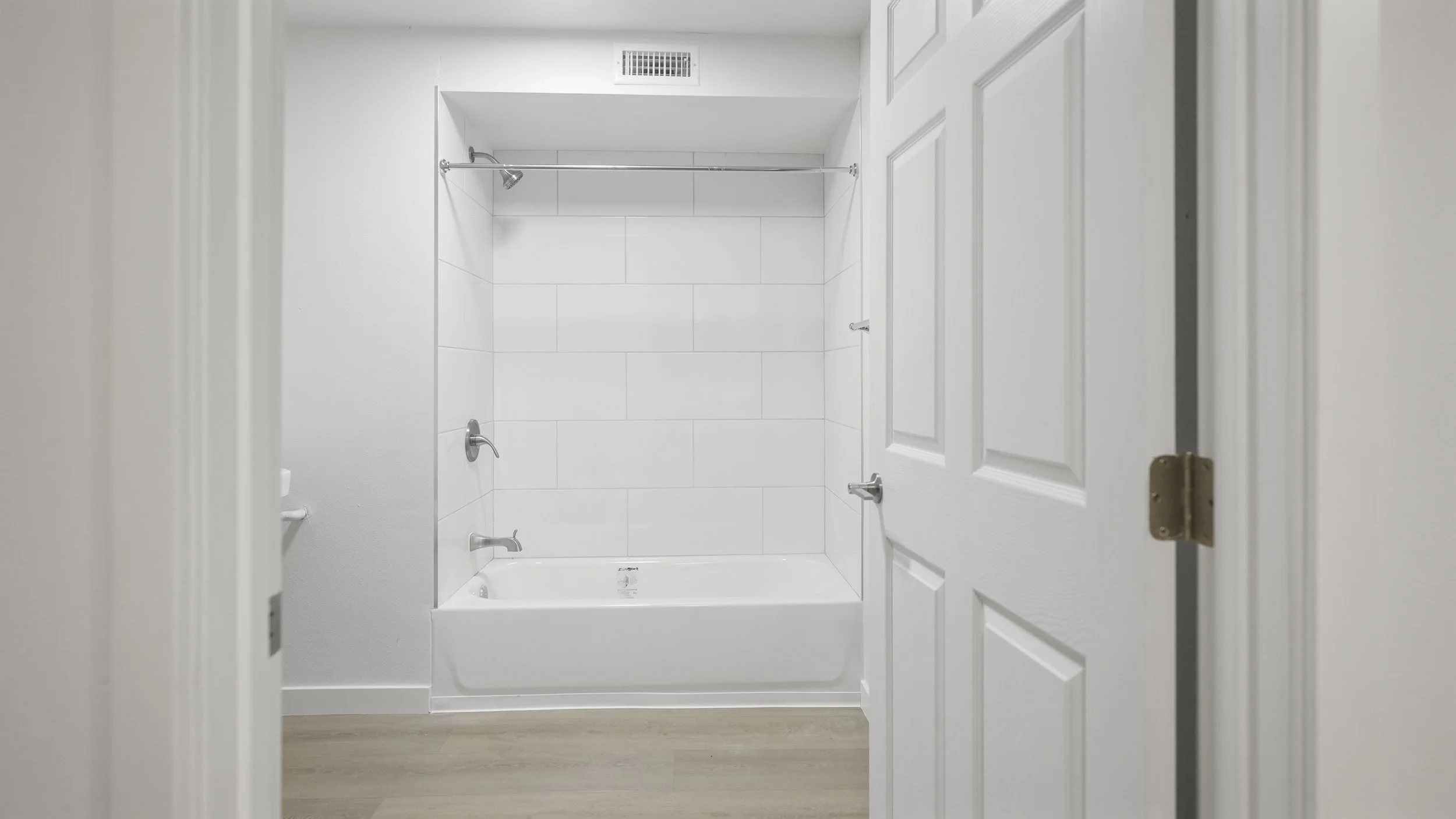 View of a clean, white bathroom with a bathtub, situated behind a partially open white door with decorative paneling; the walls and tiles are white, and there is a silver showerhead and faucet in the bathtub area.