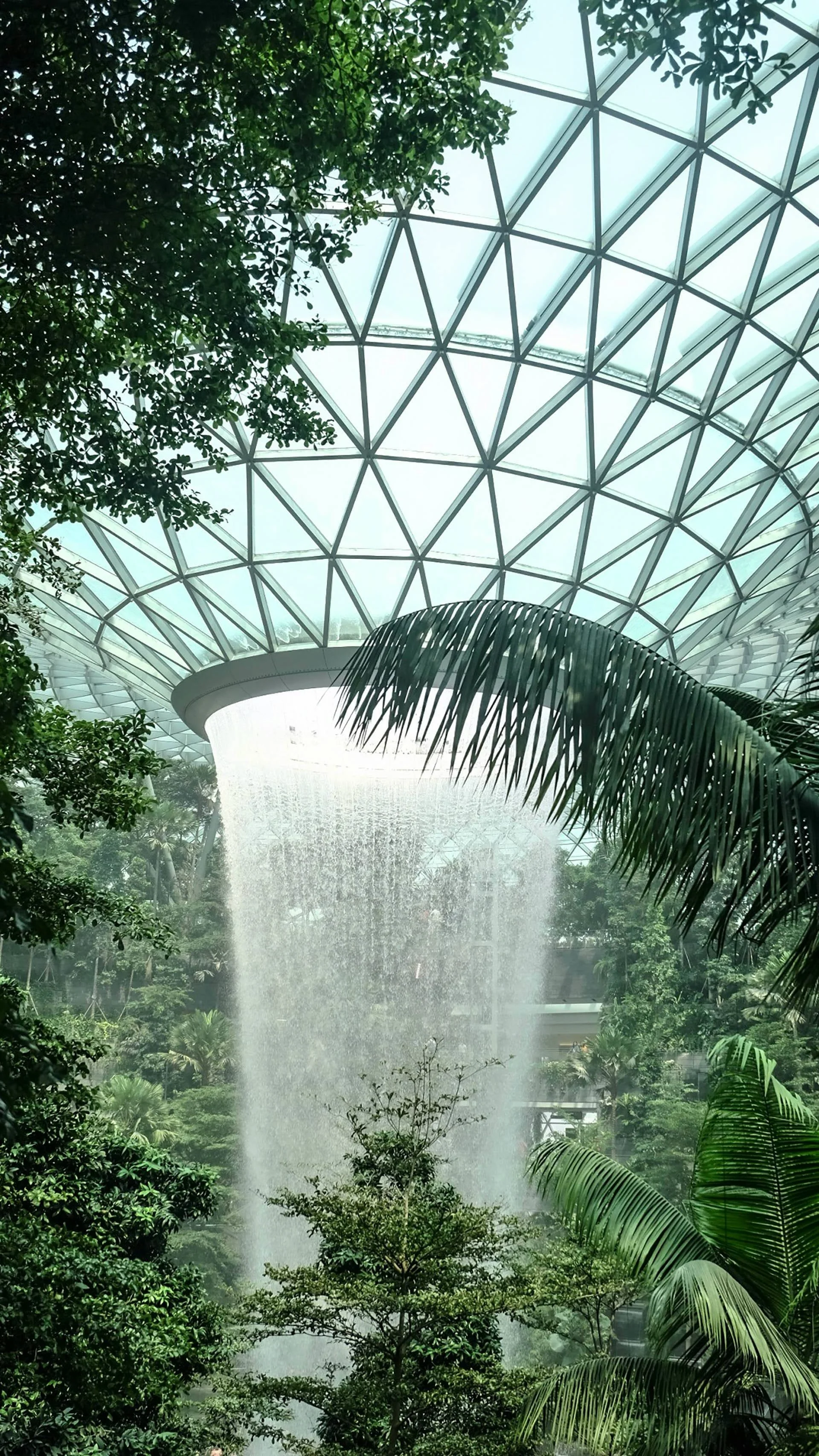 Indoor tropical garden with lush green plants and trees underneath a large geodesic glass roof, featuring a waterfall cascading from a circular opening in the ceiling.
