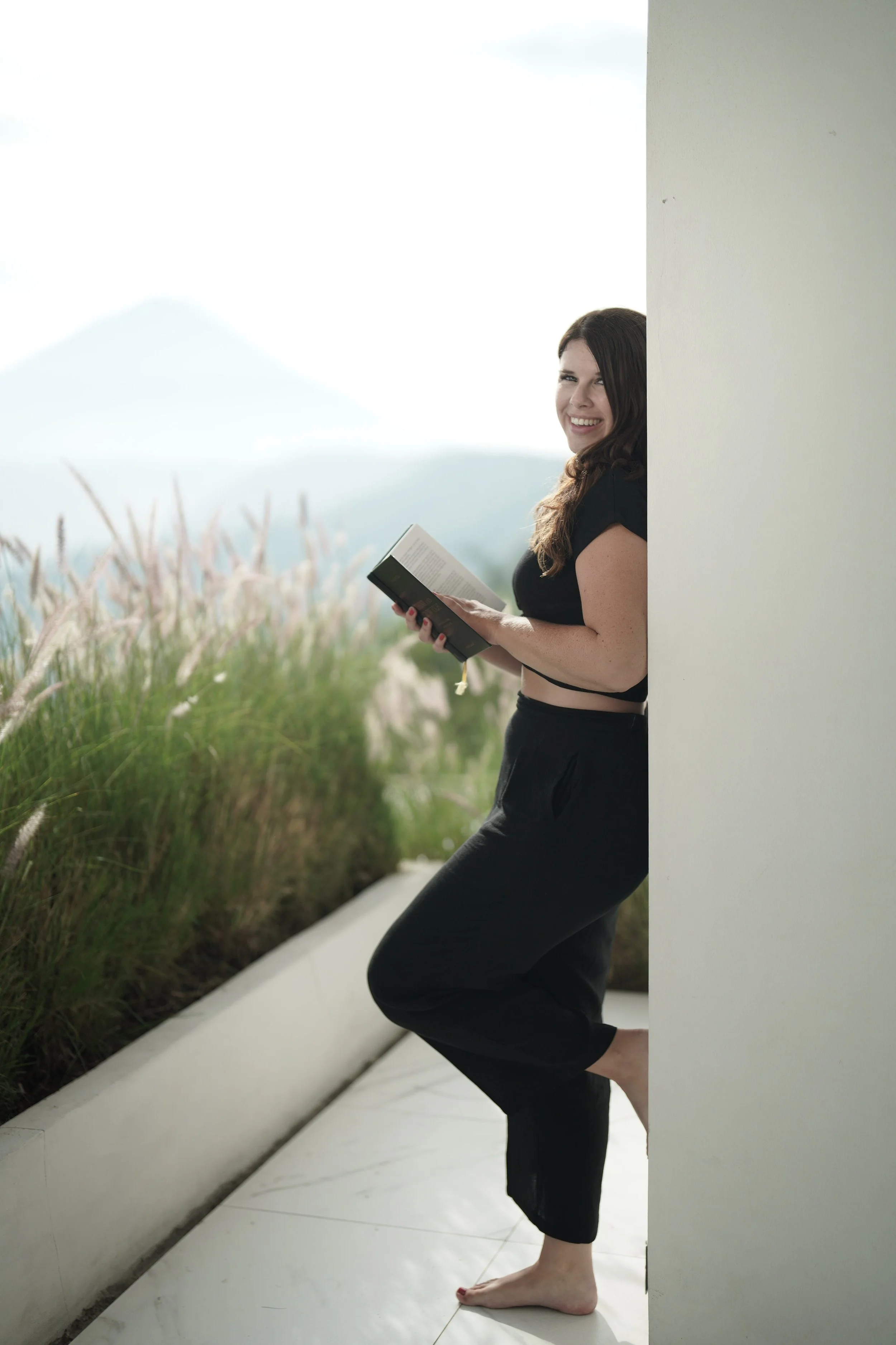A woman with dark hair leaning against a white wall, smiling, holding a book, standing barefoot on a white surface, with tall grass and mountains in the background.