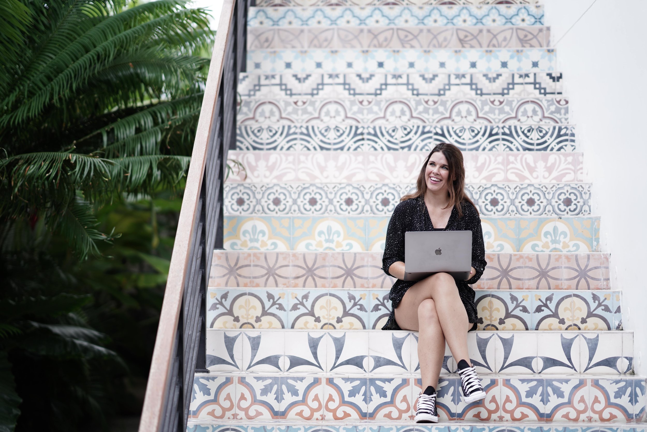 A woman sitting on colorful tiled stairs with a laptop, smiling, next to green plants.