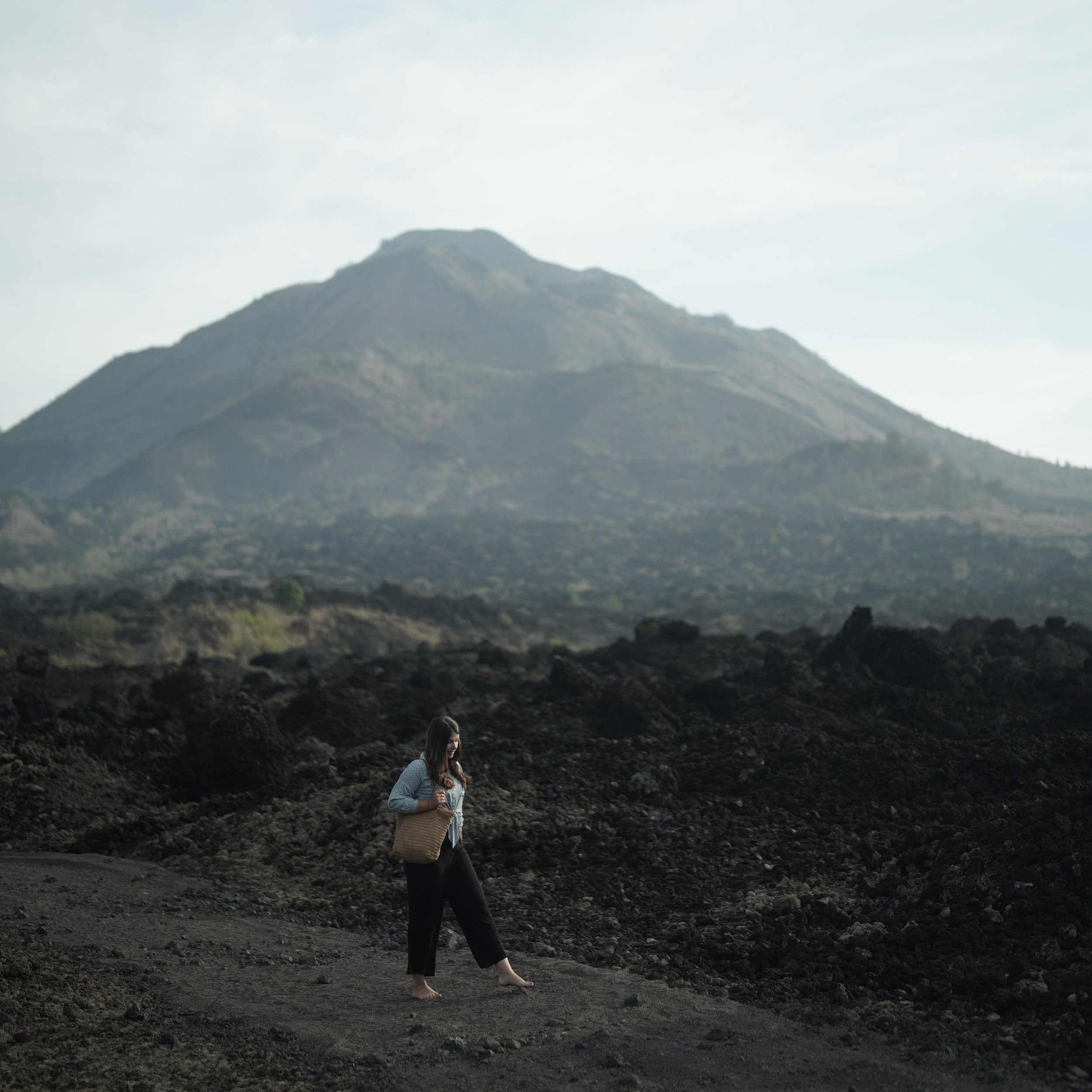 A woman walking barefoot on a rocky volcanic landscape with a large mountain in the background.